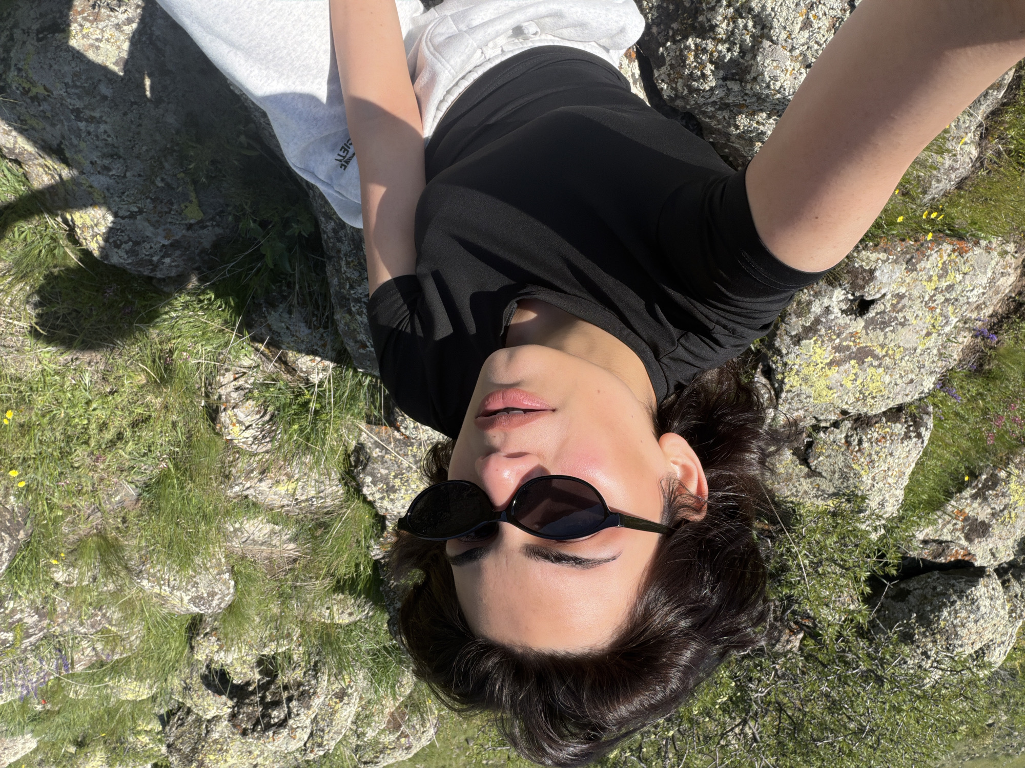 The image shows a person lying on a rocky, grassy surface in what appears to be an outdoor setting. The person is wearing sunglasses, a black shirt, and gray sweatpants. The background consists of rocks covered with lichen and grass, with some small yellow and purple wildflowers visible. Based on the information provided, this location is Tlik, Armenia. The person appears to be taking a selfie, as their arm is extended towards the camera.