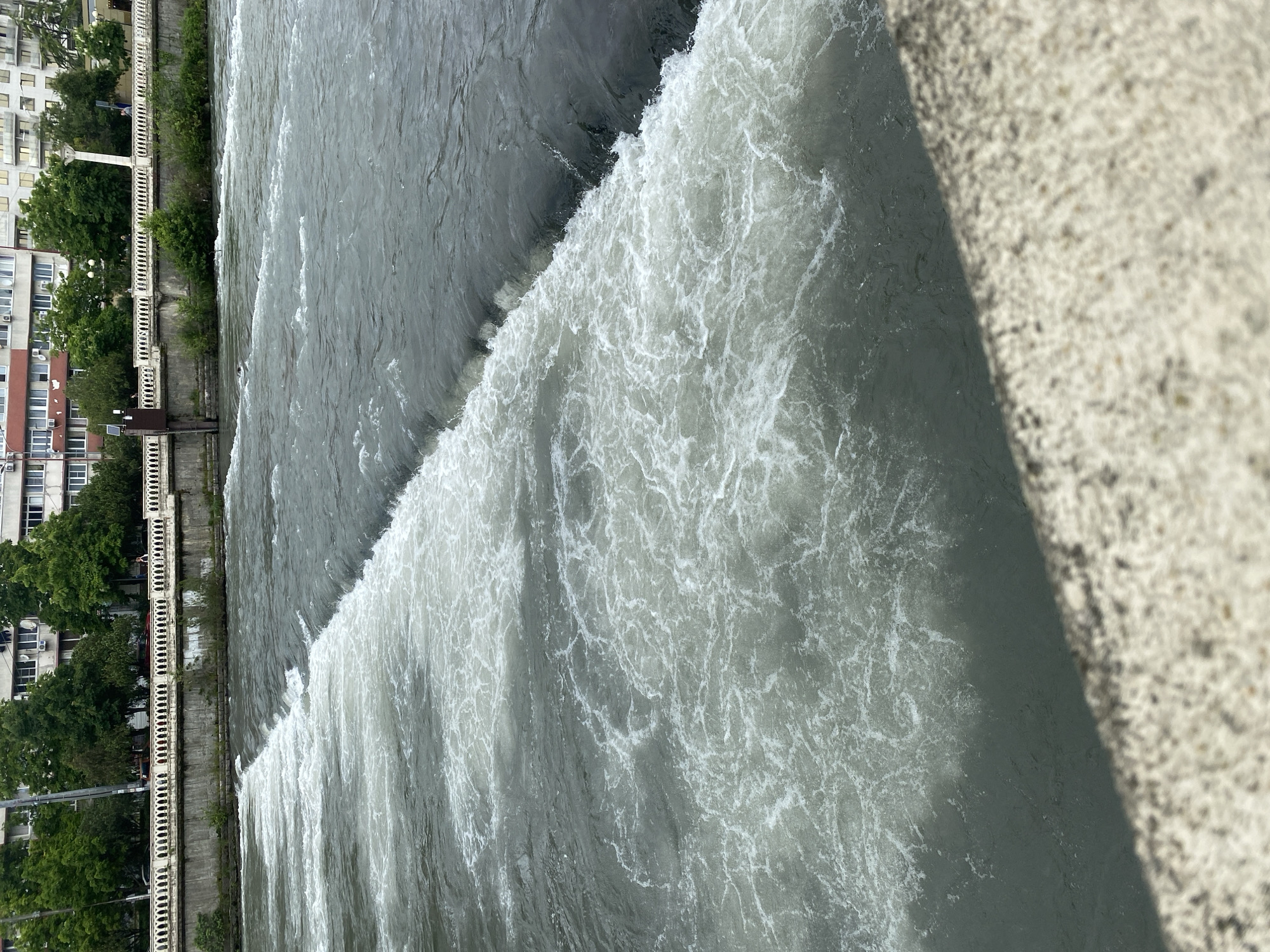 The image shows a river in Sochi, Russia. Water is cascading over a man-made weir in the foreground, creating a foamy texture on the surface. Beyond the weir, the river flows towards the left side of the frame. The embankment is visible on the left. There are buildings and trees on the embankment behind the river. A concrete wall is present in the foreground on the right side. The photo is taken from an elevated perspective.