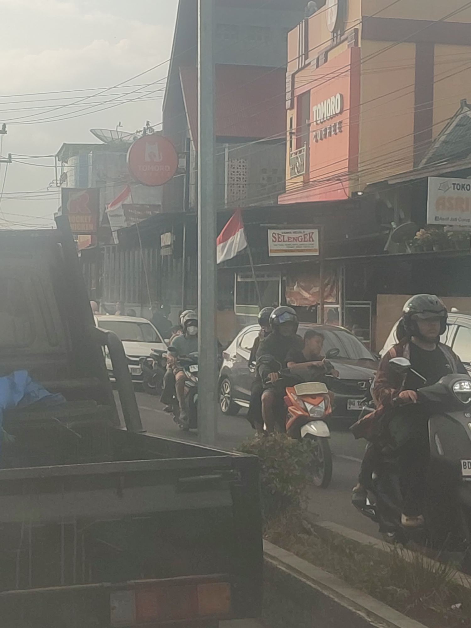 This is a medium-distance, slightly hazy, outdoor shot taken from a vehicle, likely in Adirejo, Indonesia. The scene depicts a street with moderate traffic, primarily motorcycles and cars. In the foreground, the side of a dark-colored utility truck is visible on the left. On the right side of the street, several motorcycles are present. One motorcycle in the mid-right has a rider wearing a dark helmet and a plaid jacket, with another rider behind them. In the background, buildings line the street, some featuring signage. One prominent sign reads "TOMORO COFFEE" in large, white letters against an orange building. Another sign below it says "SELENGEK". Indonesian flags are visible, with red and white colors displayed on banners and possibly on a motorcycle. The overall atmosphere suggests a typical day in a busy urban or semi-urban environment.