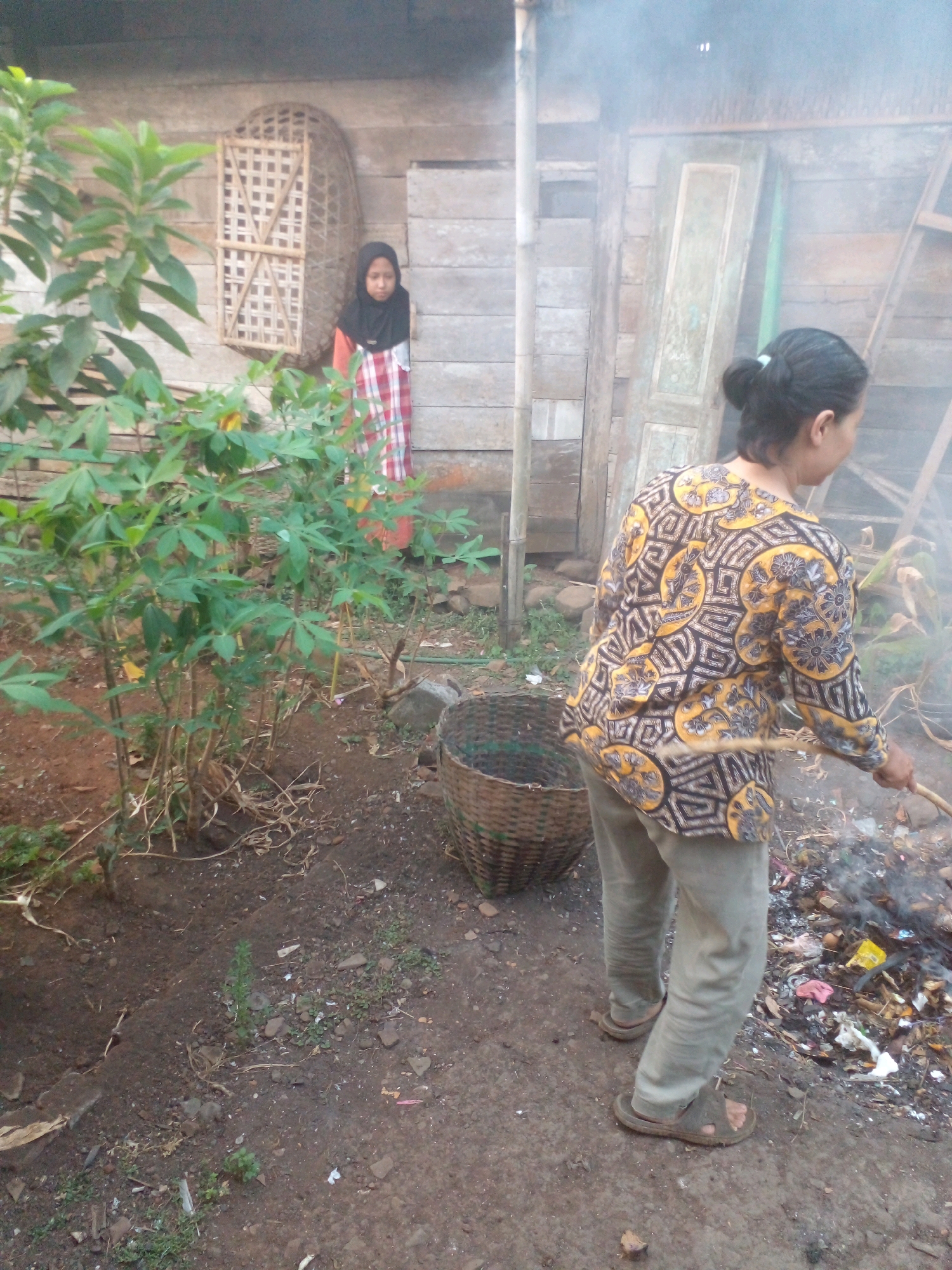 In the village of Pungangan, Indonesia, a scene unfolds in a rustic outdoor setting. In the foreground, a woman, her back turned to the viewer, wears a long-sleeved patterned shirt and light-colored pants. She stands near a pile of burning debris, actively tending to it with a stick. To her left, a woven basket sits on the dirt ground. Behind her, a young girl in a hijab and a checkered dress stands near a wooden structure with a decorative woven element. Lush green plants with large leaves dominate the left side of the frame. The environment appears to be a yard or open space adjacent to a wooden building.