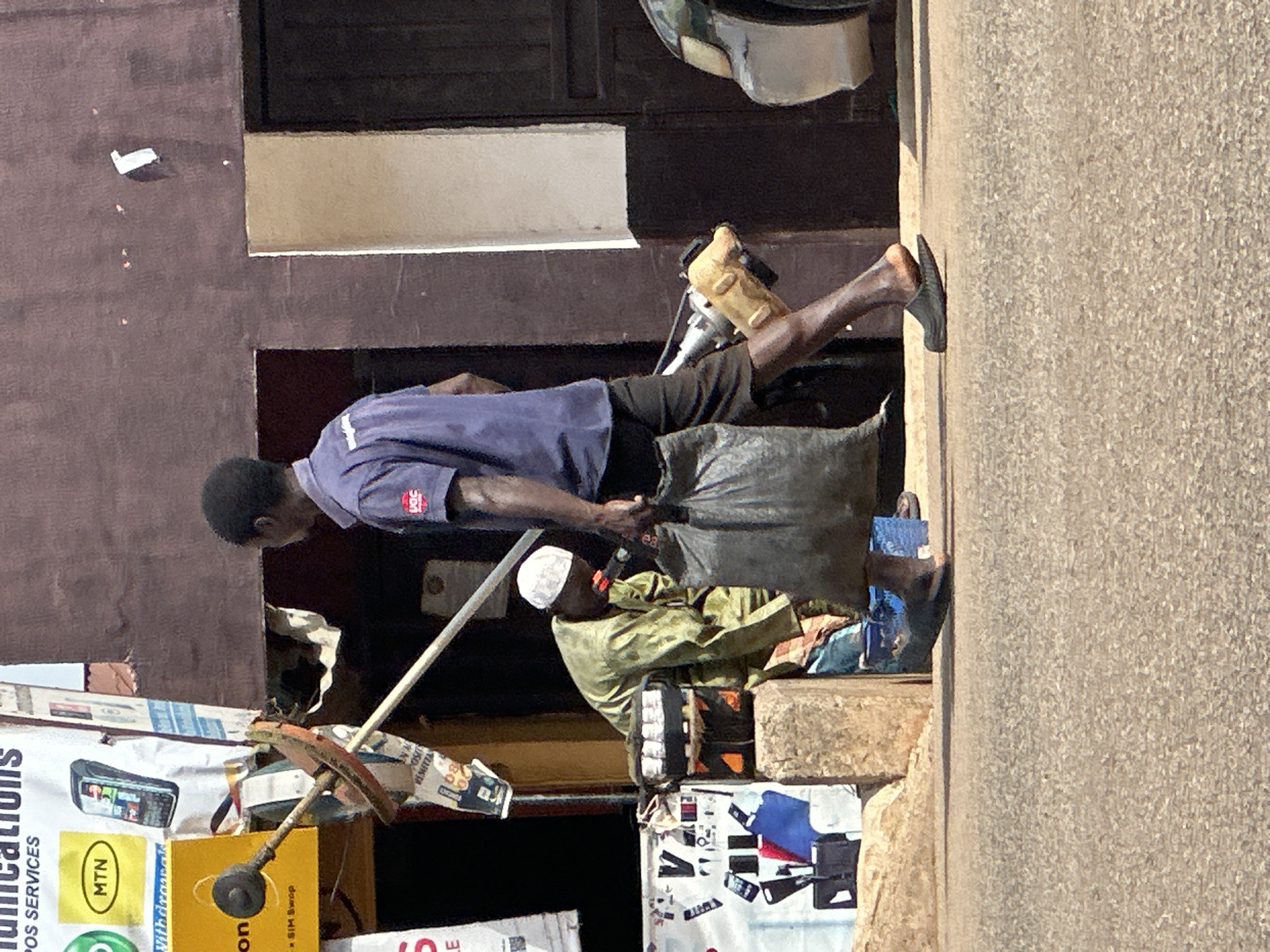 In Bukuru, Nigeria, a man walks past a stall. The stall displays various telecommunication services, including MTN, phone chargers, and SIM swap services. The man is wearing a purple t-shirt and brown shorts, carrying a large black bag over his shoulder. To his left, another man in a green traditional outfit and a white cap is attending to a POS machine. The scene is set on a paved street with a building in the background.