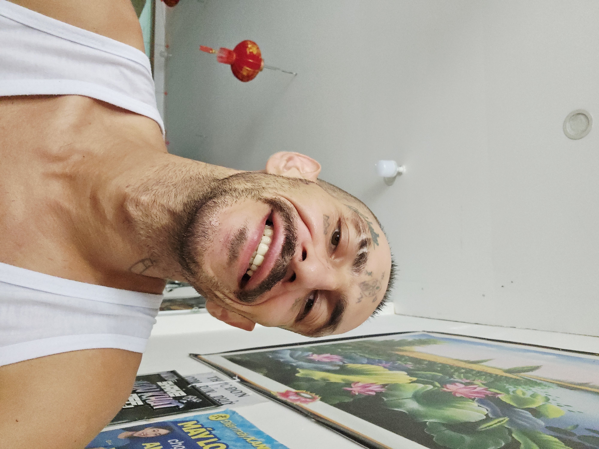 A man with a beard and visible tattoos is smiling broadly and looking at the camera. He is wearing a white tank top. A framed picture depicting lotus flowers and a blue sky is on a white surface in the foreground. There are advertisements and magazines, one with the word "MÁYLO" visible, next to the framed picture. A red lantern is visible hanging from the ceiling in the background. The image may have been taken in Cam Ranh Ward, Vietnam, based on the language of the advertisement. The room appears to be indoors, with white walls and a light fixture.