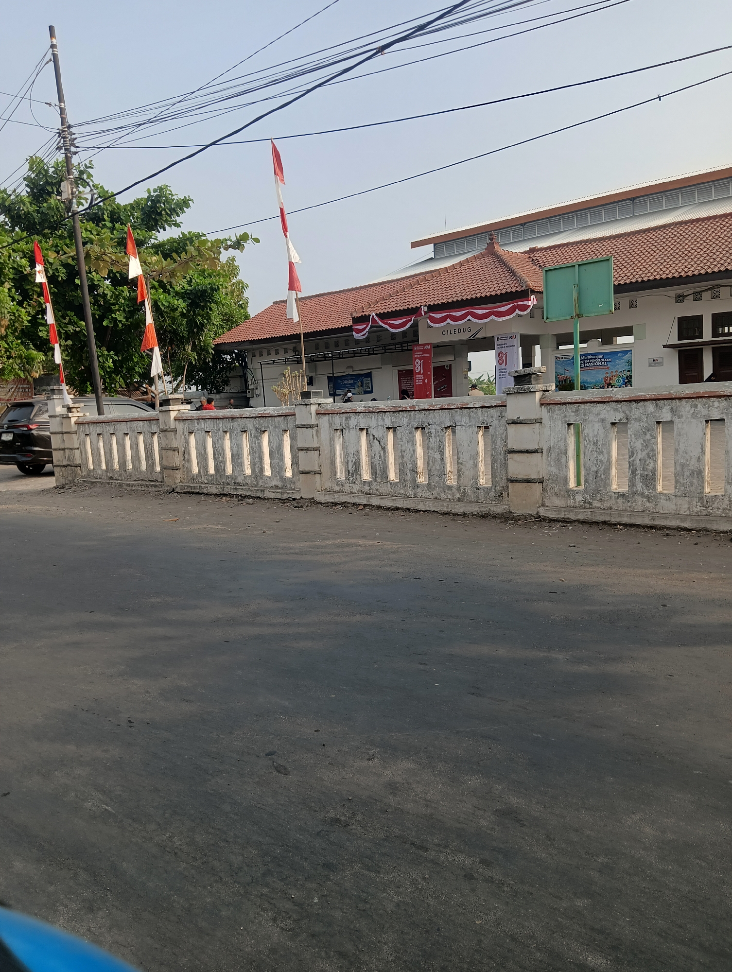 The image depicts a street scene in front of a building identified by signage as "CILEDUG." The building has a red tile roof and white walls. In front of the building is a low, light-colored concrete fence with rectangular openings. Several Indonesian flags, red and white, are displayed on poles. A dark-colored vehicle is visible on the street to the left of the frame. The environment appears to be outdoors, with a clear sky and some trees visible. The street in the foreground is paved with asphalt. There are power lines visible above the building.