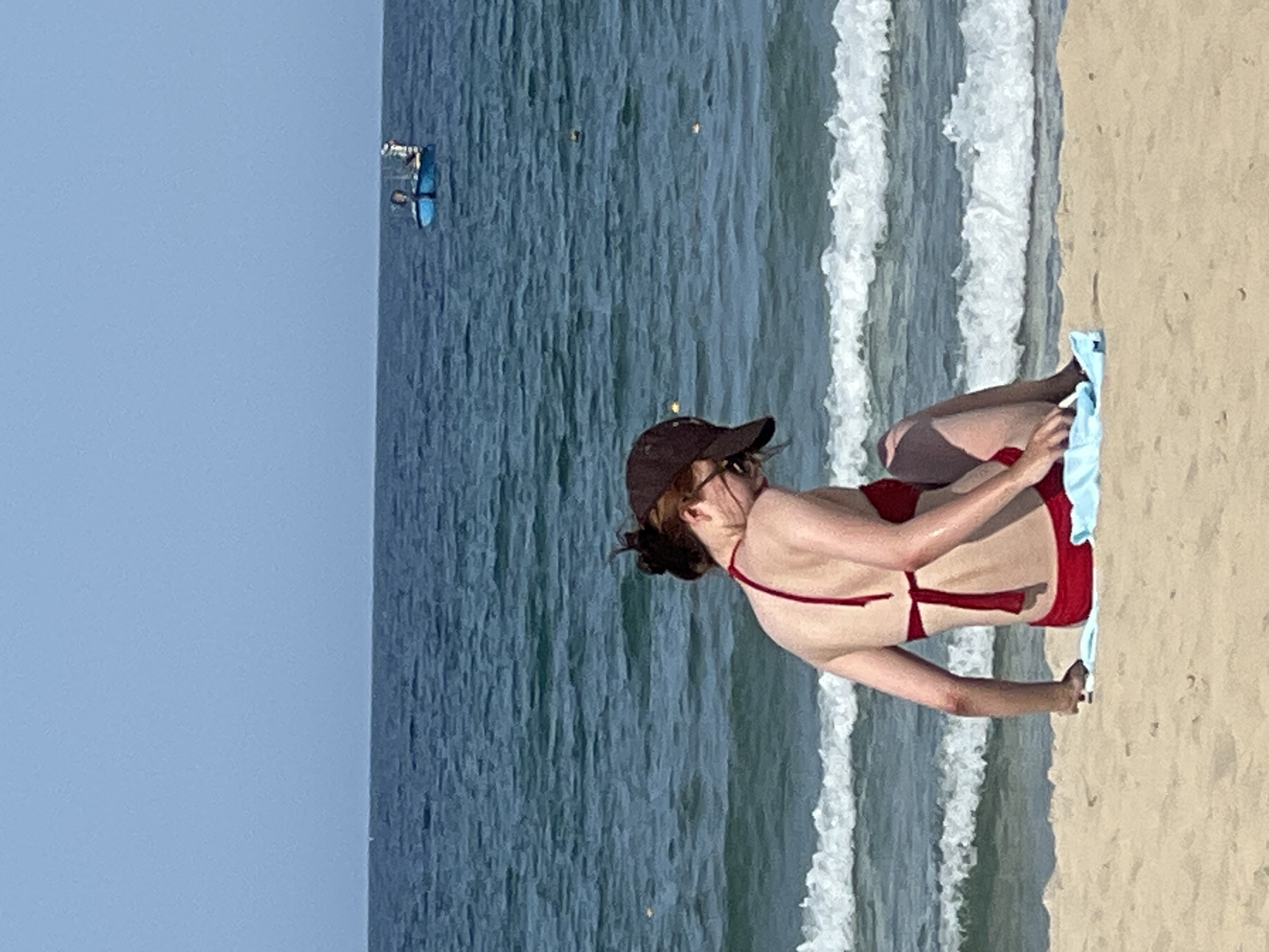 The image depicts a beach scene in Da Nang, Vietnam. In the foreground, a person wearing a red bikini and a dark baseball cap is sitting on a light blue towel on the sand. The person is facing away from the camera, with their head tilted down. The person's left arm is resting on their lap, and their right hand is on the sand. The sea is visible in the background, with gentle waves breaking on the shore. A small blue boat with two figures is present in the distance on the water, under a clear blue sky. The sand appears light brown and undisturbed, with small ripples from the receding waves.