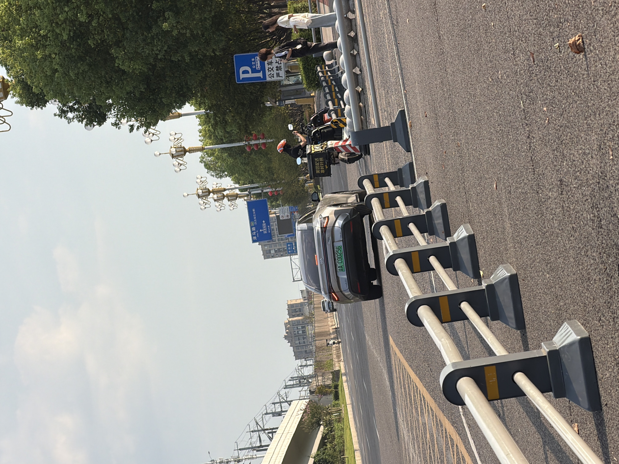 An outdoor, eye-level, medium shot captures a street scene in Jiangjin, China, under a bright sky with scattered clouds. In the foreground, a metal and plastic barrier runs along the edge of an asphalt road. The road has yellow and white lane markings. Further down the road, a silver sedan with a green license plate displaying "A C00256" is traveling away from the camera. To the right of the sedan, a person wearing a red helmet and black clothing is stopped on a scooter with a large yellow delivery box attached. The scooter is partially obscured by a traffic cone with red and white stripes. Two individuals stand on the sidewalk to the right, one wearing a black jacket and the other a white top and grey pants. A blue and white "P" sign with a bus symbol and Chinese characters indicates a bus stop. Streetlights with decorative gold elements line the road, and lush green trees are visible behind them. In the background, a large, elevated concrete structure, possibly an overpass or bridge, is visible, with buildings and overhead railway lines stretching into the distance. The overall impression is a typical urban street scene on a clear day.