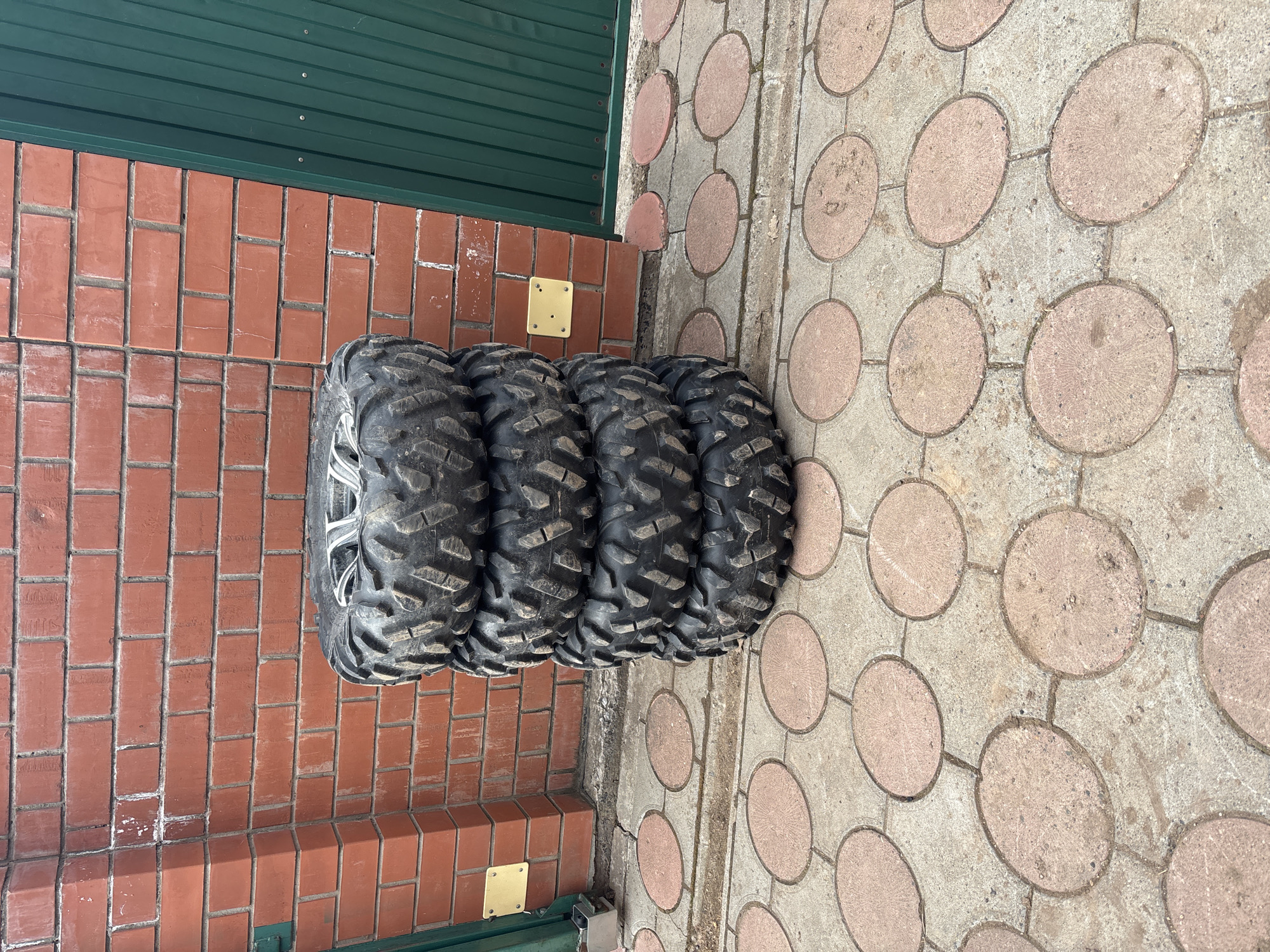 Four off-road tires are stacked against a brick wall in Ферма, Russia. The wall is made of red bricks and is partially covered by a green metal panel at the top. A yellow mounting plate is visible on the wall near the base. The tires are positioned on a paved surface with a pattern of large circles embedded in concrete.