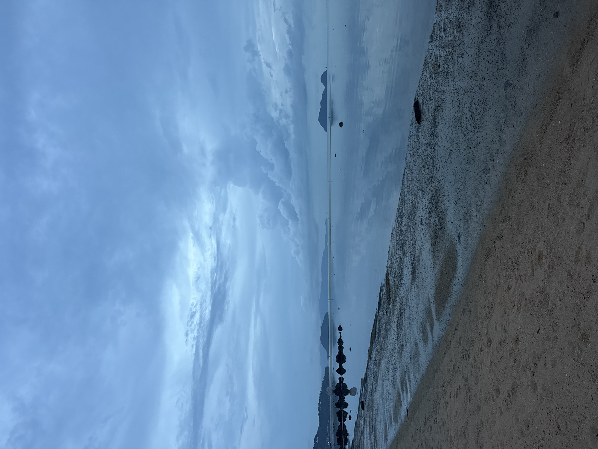 The scene presents a view of a beach in Baan Sri Thanu, Thailand. The sky is cloudy, with visible cloud formations above water with mountainous land masses in the distance. The foreground features a sandy beach area, partially wet from the receding water line, on the lower right section of the image.