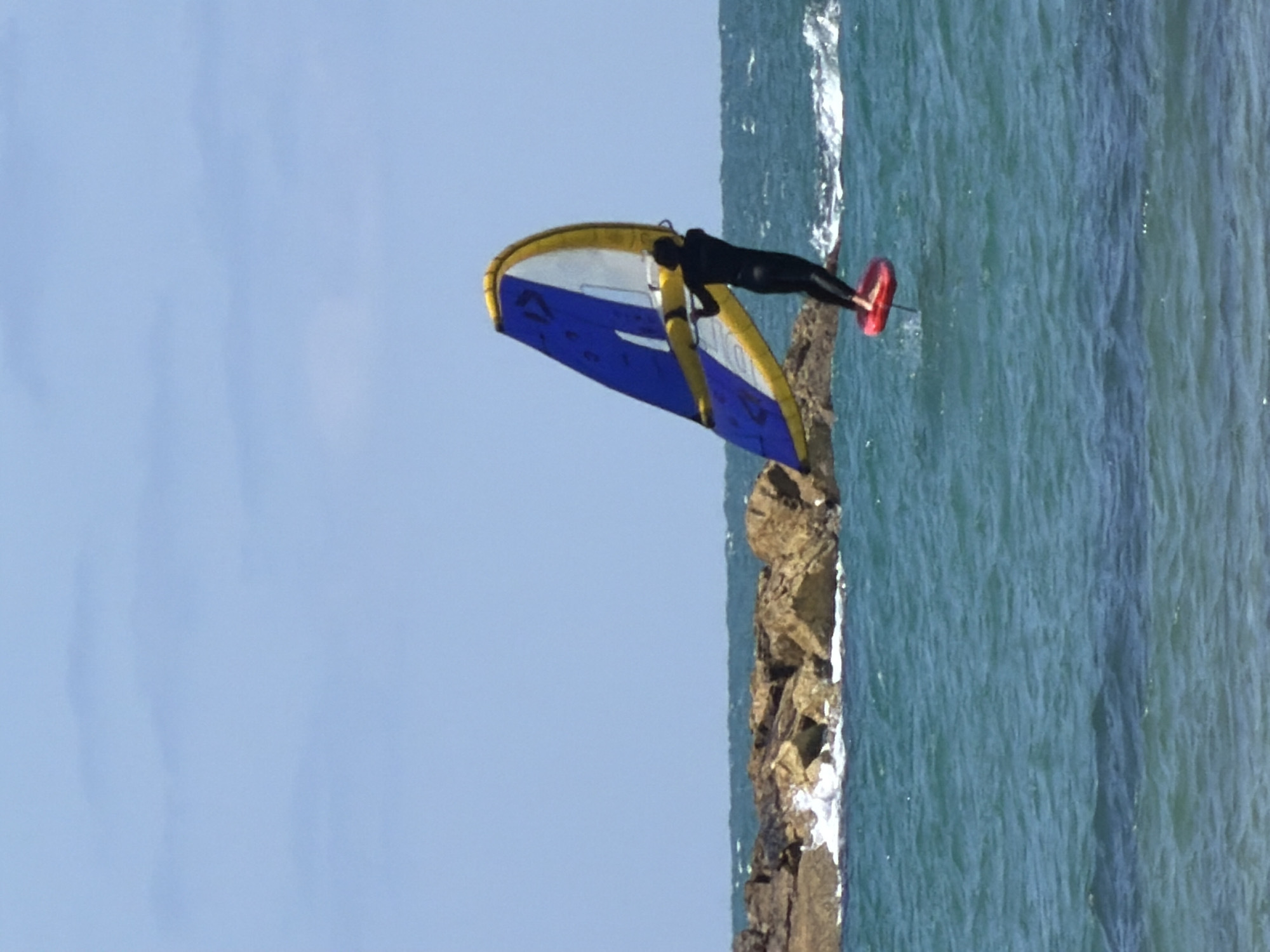 A person is wing foiling off the coast of Tel-Aviv, Israel. The person is wearing a black wetsuit and is holding onto a blue and yellow wing. They are riding on a hydrofoil board with a red leash. The water is a greenish-blue color, and there are some rocks visible along the shoreline. The sky is pale blue.