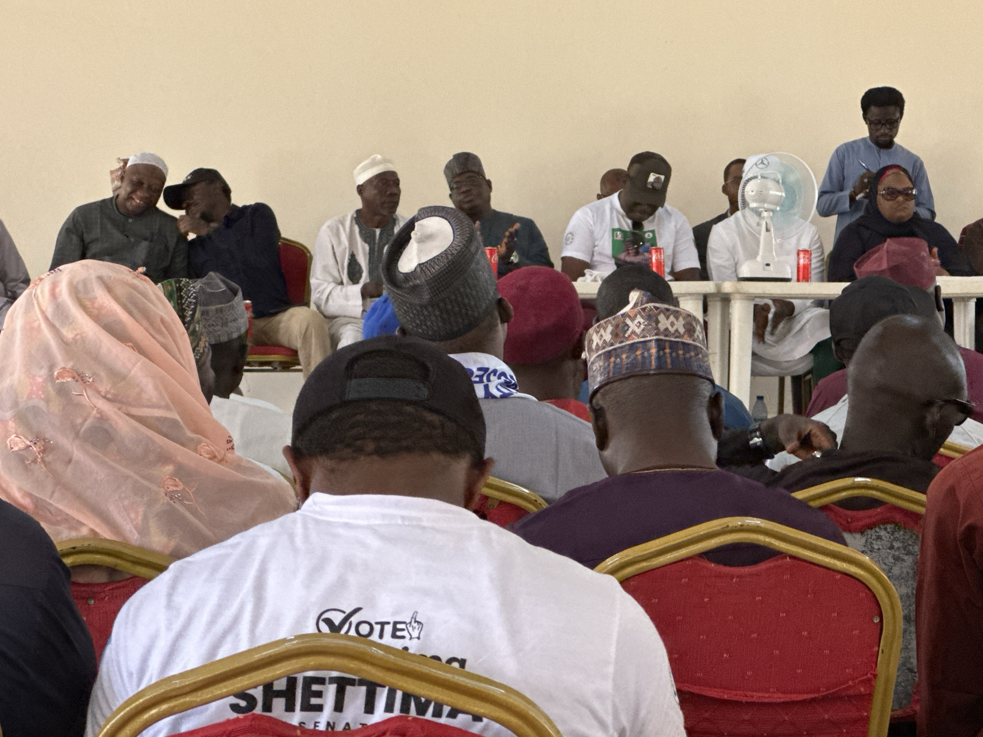 A medium shot shows a gathering of mostly adult men in an indoor space in an unknown city. They are seated in chairs, presumably attending a meeting or event. Some are dressed in traditional Muslim attire, including various styles of caps and robes. Others wear casual clothing such as t-shirts. Notably, one t-shirt in the foreground displays "Vote SHEITTIMA SENATORIAL." In the background, a fan stands on a table. The walls are a plain, neutral color, and lighting is diffused. The specific location within the city is not discernible.