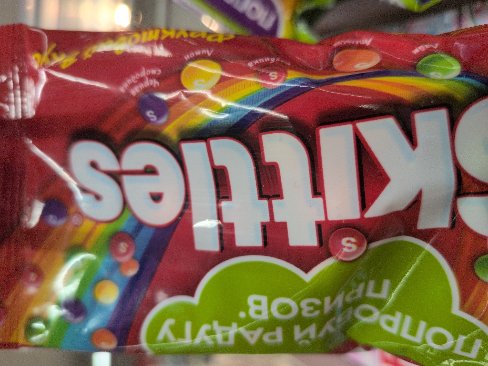 The image provides a close-up view of a Skittles candy package, featuring a red background with a vibrant rainbow stripe and illustrations of colorful candy pieces. The prominent "Skittles" brand name is visible in large white letters across the center of the package, appearing mirrored or reversed in the image.
Above the rainbow, various Cyrillic flavor names are printed next to their corresponding candy colors, also reversed. These include "черная смородина" (blackcurrant) for purple, "лимон" (lemon) for yellow, "клубника" (strawberry) for red, "апельсин" (orange) for orange, and "лайм" (lime) for green. Each depicted candy piece features a small white "S" logo.
At the bottom of the package, a green, cloud-like graphic contains further Cyrillic text, which is both reversed and upside down. It reads "ПОПРОБУЙ РАДУГУ ВКУСОВ", translating to "TASTE THE RAINBOW OF FLAVORS."
The packaging exhibits some creases and reflections from bright, artificial light sources, indicating an indoor setting, likely a retail environment. No people are present. The use of Cyrillic text confirms the product is marketed in a Russian-speaking region, aligning with the provided location context of Sochi, Russia. The time of day is not discernible.