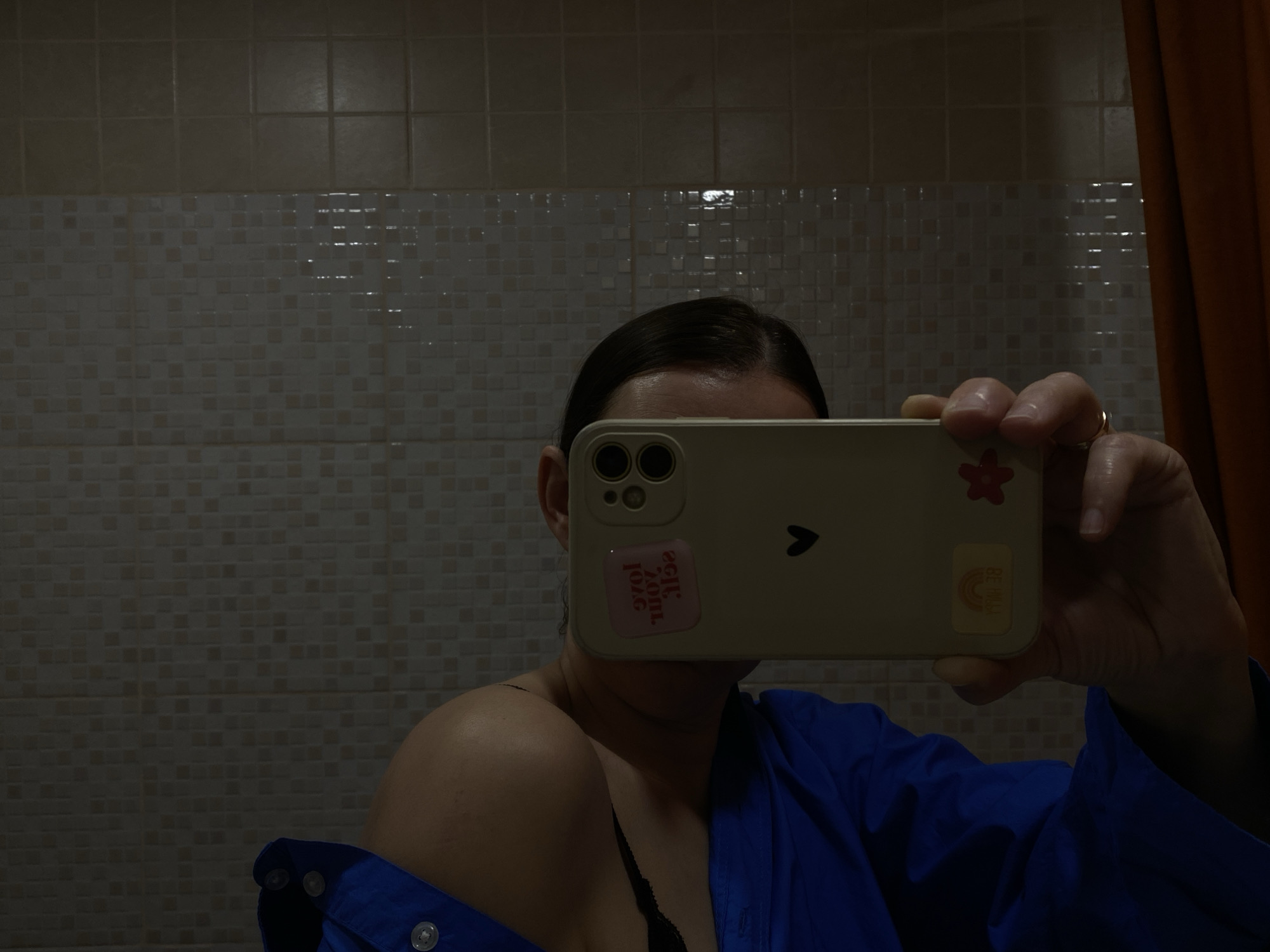 A person is taking a selfie in a bathroom. The person is holding a smartphone in front of their face, obscuring most of it. The phone has stickers on it. The person is wearing a blue shirt or robe, partially open to reveal a black bra strap. The background shows tiled walls and an orange curtain. The exact location in Chaikovsky, Russia, is unknown, but it is a bathroom.