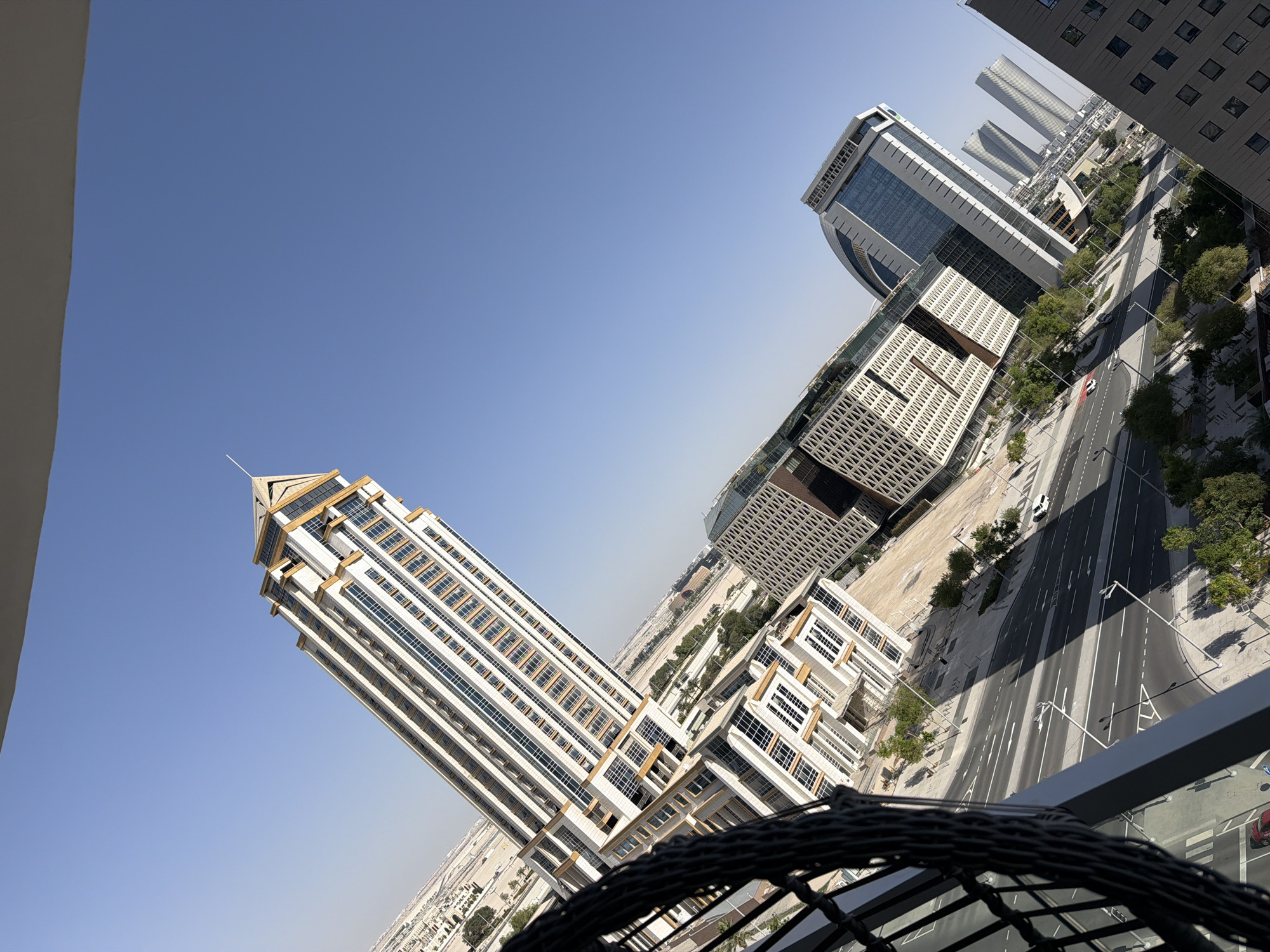 A high-angle, tilted shot captures a modern cityscape under a clear blue sky. In the foreground, a dark, woven railing obscures the lower left corner. The primary focus is a cluster of contemporary buildings. Dominating the left side is a tall, slender skyscraper with a peaked, golden-accented roof and many windows. To its right, a lower, elongated building features a patterned facade and a glass-fronted section. Further to the right, a curved, glass-clad building stands out, with three similarly shaped, ribbed towers visible in the background. A multi-lane road runs through the scene, with white lane markings and a few scattered vehicles, including a white car. Trees with green foliage line the road and are interspersed between the buildings. The ground surrounding the buildings appears to be paved or constructed. The overall impression is one of urban development and modern architecture.