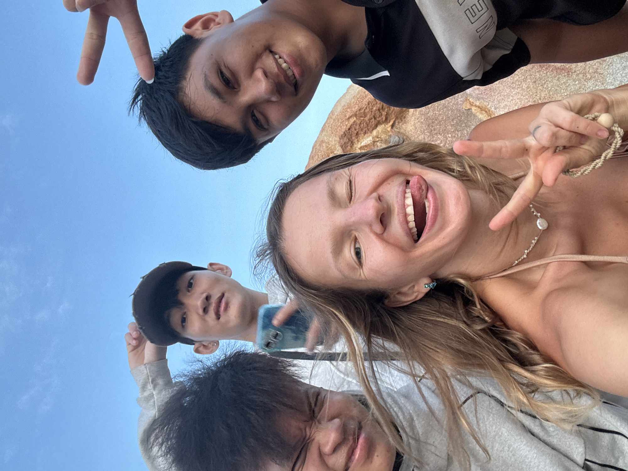 A group of four young people are taking a selfie outdoors under a clear blue sky. In the foreground, a blonde woman is winking and sticking her tongue out, making a peace sign with her right hand. To her left, a young man with dark hair is making a disgusted face. Behind them, another young man in a dark cap holds up a phone. To the right of the woman, another young man in a black polo shirt smiles at the camera. The background shows a large rock and the sky. The image appears to have been taken on a beach or coastal area.