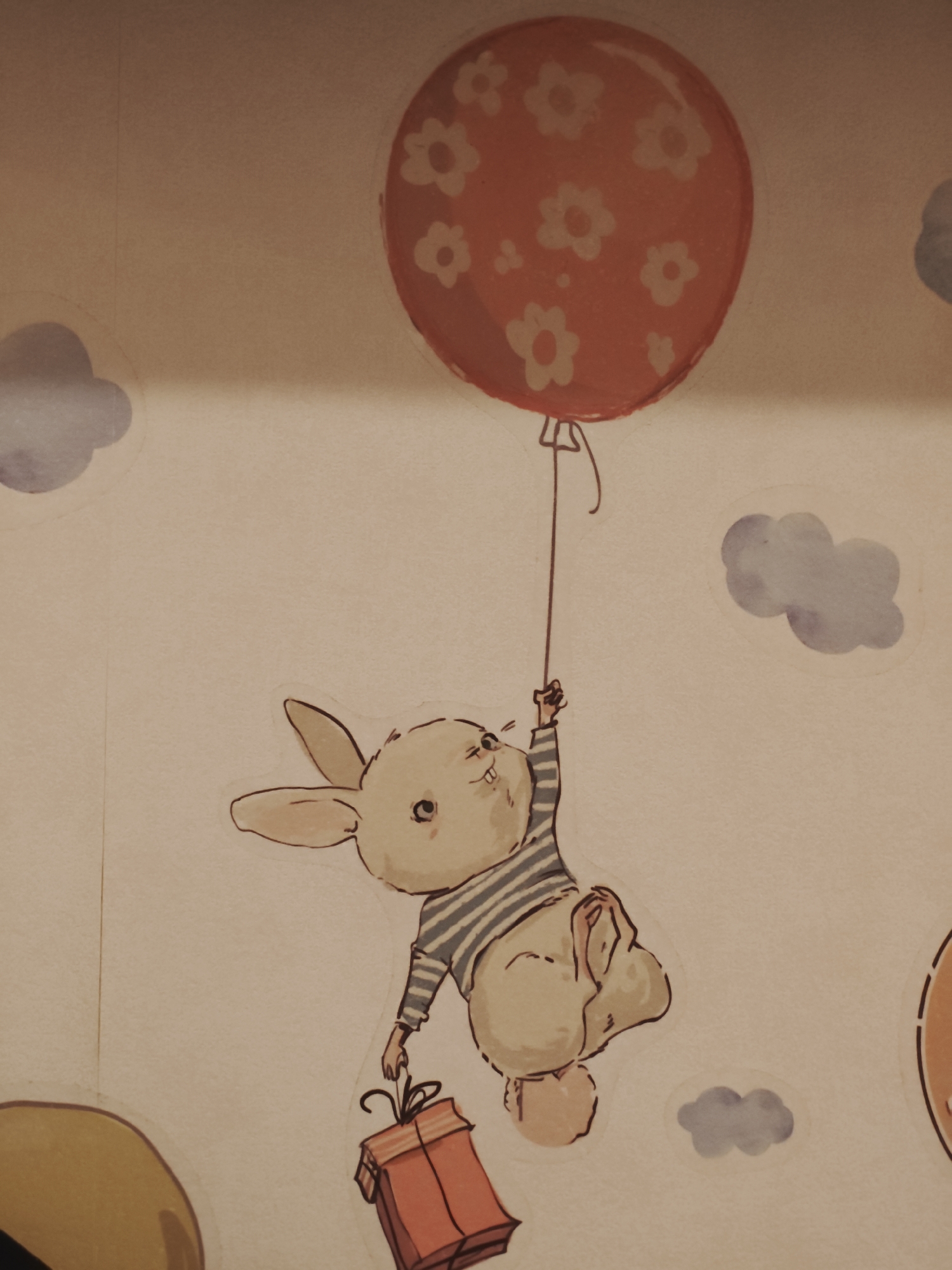 The image shows a drawing on a light-colored, textured surface, possibly wallpaper or a canvas. The drawing depicts a stylized rabbit character holding onto a balloon string, which is attached to a large, red balloon decorated with white flowers. The rabbit is wearing a striped shirt and appears to be floating. It is also holding a small, orange gift box with a ribbon. Several small, blue watercolor-style clouds are scattered around the scene, suggesting a sky setting. The overall style of the drawing is whimsical and childlike. There is no indication of this image being located in Saint Petersburg, Russia; the image only depicts a drawing.