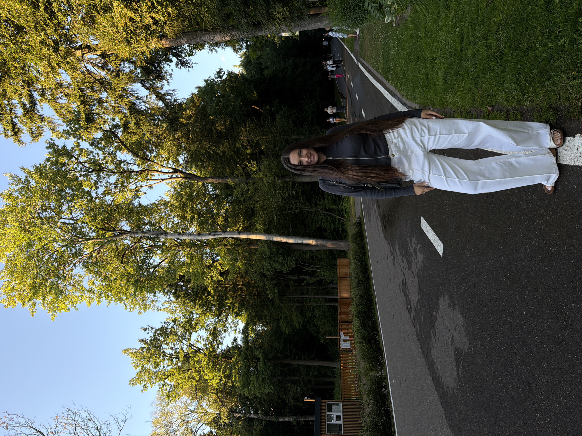 A woman with long, dark hair stands on an asphalt path in a park. She is wearing a dark gray zip-up jacket and wide-leg white pants. She is facing the camera and smiling. The path is lined with green grass and tall trees. In the background, there are several other people walking and a wooden fence with a small building behind it. The image appears to have been taken in Odintsovo, Russia.