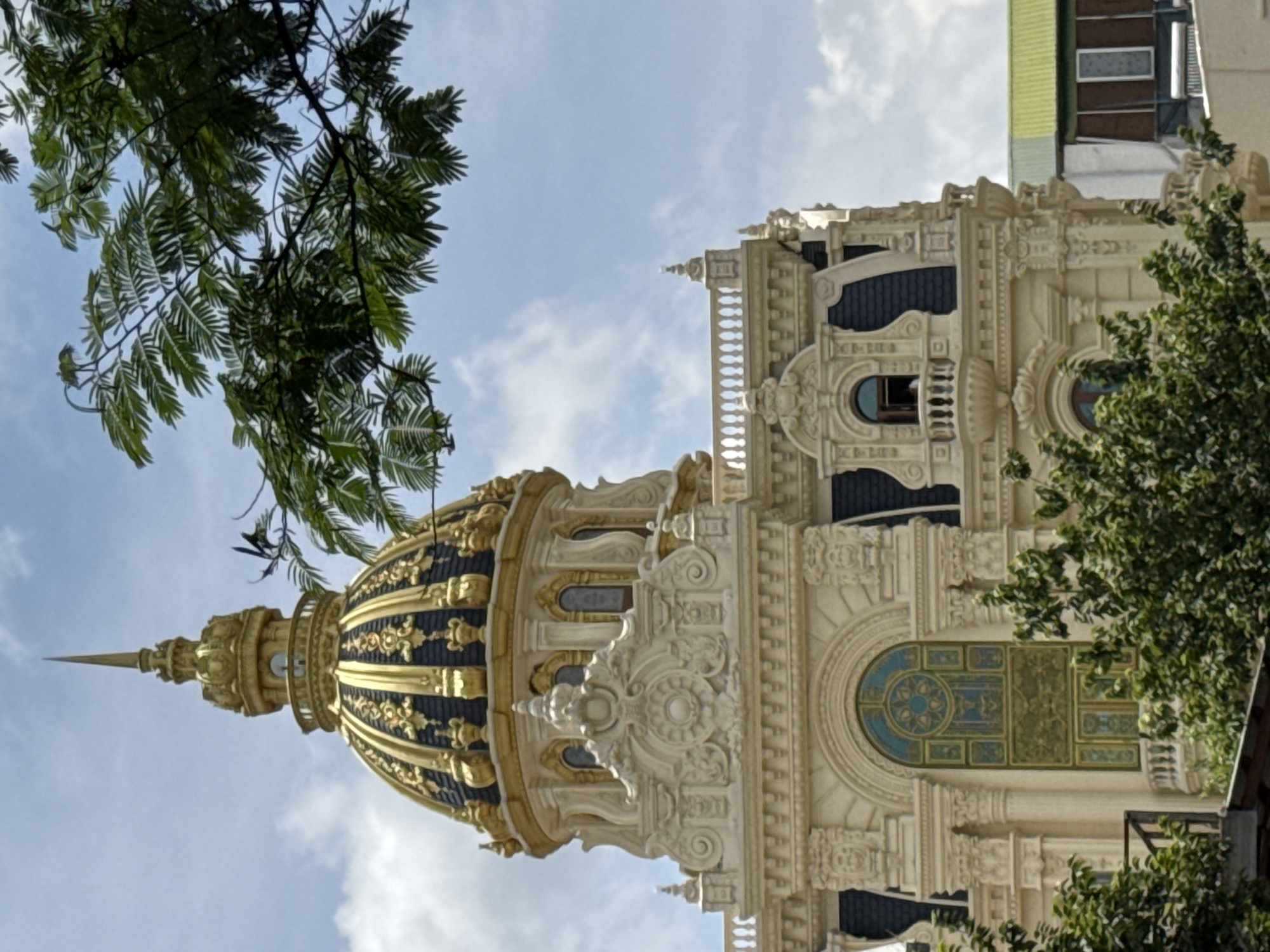 This image depicts a close-up view of a striking building in Da Lat, Vietnam, characterized by its ornate architecture. The building features a prominent golden dome topped with a spire, adorned with intricate dark patterns and decorative elements. The facade is richly decorated with elaborate moldings, carvings, and ornate windows. In the foreground, lush green trees partially obscure the lower portions of the building, suggesting an outdoor setting. The sky above is blue with scattered clouds. The overall impression is one of grandeur and historical architectural style.