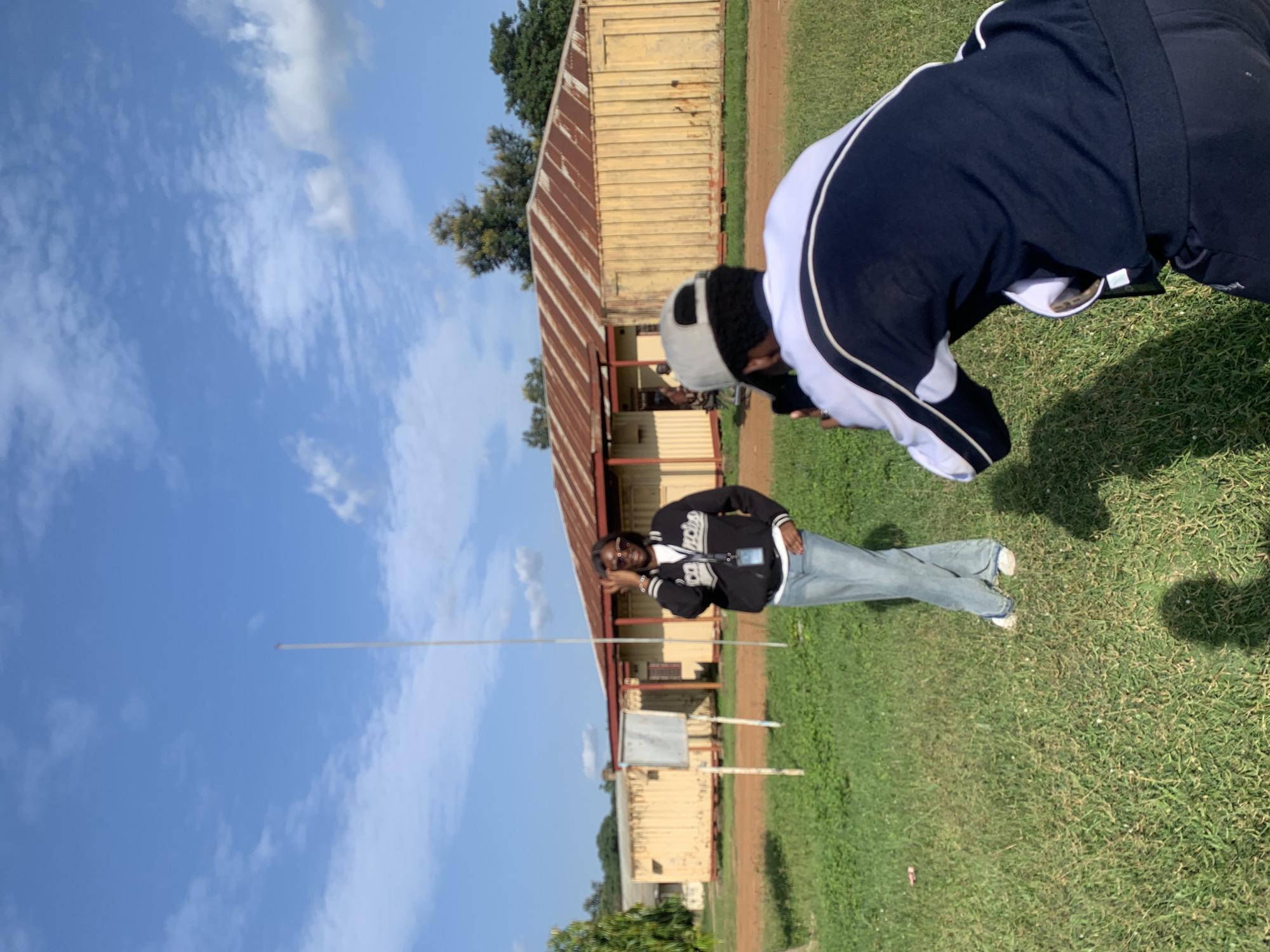 In Mangu Town, Nigeria, on a sunny day with a blue sky and scattered clouds, a person is being photographed. The photographer, wearing a grey baseball cap and a dark blue and white jacket, is positioned in the foreground, out of focus, and is kneeling while holding a phone to take a picture. The subject, a person wearing sunglasses, a black jacket with white lettering, a white shirt, and light blue jeans, is standing in the mid-ground, in focus, striking a pose with one hand near their head. Behind the subject is a building with a rusty, corrugated metal roof and yellow walls, appearing to be a classroom or administrative building. The environment is grassy with some dirt paths visible. A tall, thin white pole stands in the background.