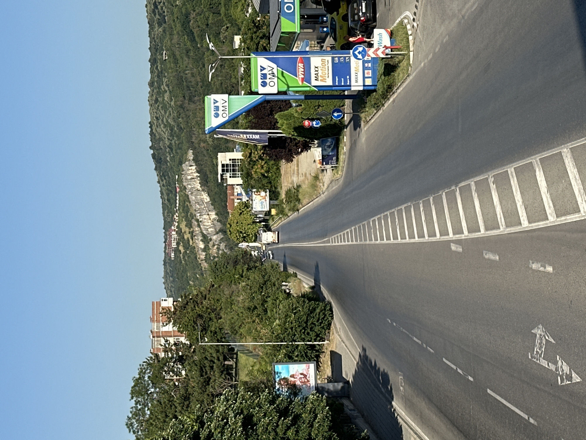 This is a photograph of a gas station on a sunny day, likely in the late morning or early afternoon, given the bright sunlight and long shadows. The scene is captured from a slightly elevated perspective, looking down a curved road. The gas station, branded with "OMV" and "VIVA MAXX Motion," is the central object in the foreground, featuring prominent signage and fuel pumps. A car is visible at the pumps, and a person in a red shirt is standing near it.
To the left of the gas station, a steep, tree-covered hillside rises. Several buildings, some with distinctive red brick accents, are nestled into the landscape at the base of the hill. The road itself is asphalt, with clear lane markings, including a dashed white line separating lanes and a solid white line along the edge. White concrete barriers with diagonal lines mark the edge of the road.
In the background, the landscape continues with more hills and greenery, suggesting a rural or semi-rural setting. The sky is a clear, bright blue. A billboard advertising an event with people is visible near the bottom of the frame, to the left of the road. The overall impression is one of a typical day at a roadside service area, surrounded by natural scenery. The text visible includes brand names like "OMV," "VIVA," and "MAXX Motion," as well as traffic signs and prices (e.g., "0.6," "15"). There's also a sign indicating "Wash."