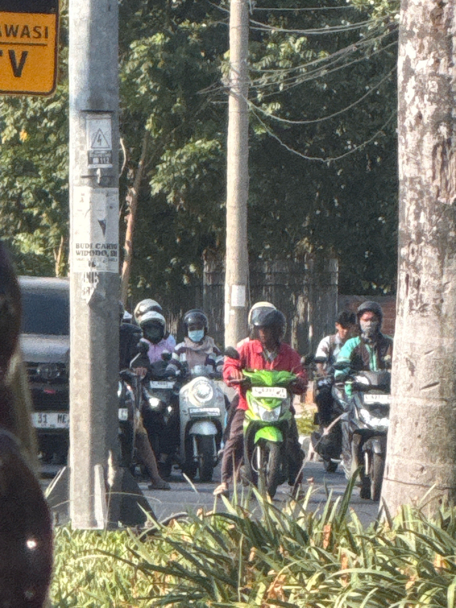 A group of motorcyclists are seen on a street in Surabaya, Indonesia, on a sunny day. The scene is bustling with activity, with several individuals wearing helmets and riding motorcycles. The foreground is dominated by lush green foliage, possibly a roadside garden or park. In the background, trees and utility poles create a typical urban landscape. One of the motorcyclists, dressed in a red jacket, is prominently featured in the center of the frame, riding a green scooter. Another motorcyclist, wearing a blue shirt and a helmet, is also visible, adding to the dynamic feel of the image. The overall impression is one of a busy street in a tropical city, with a focus on the everyday lives of its residents.