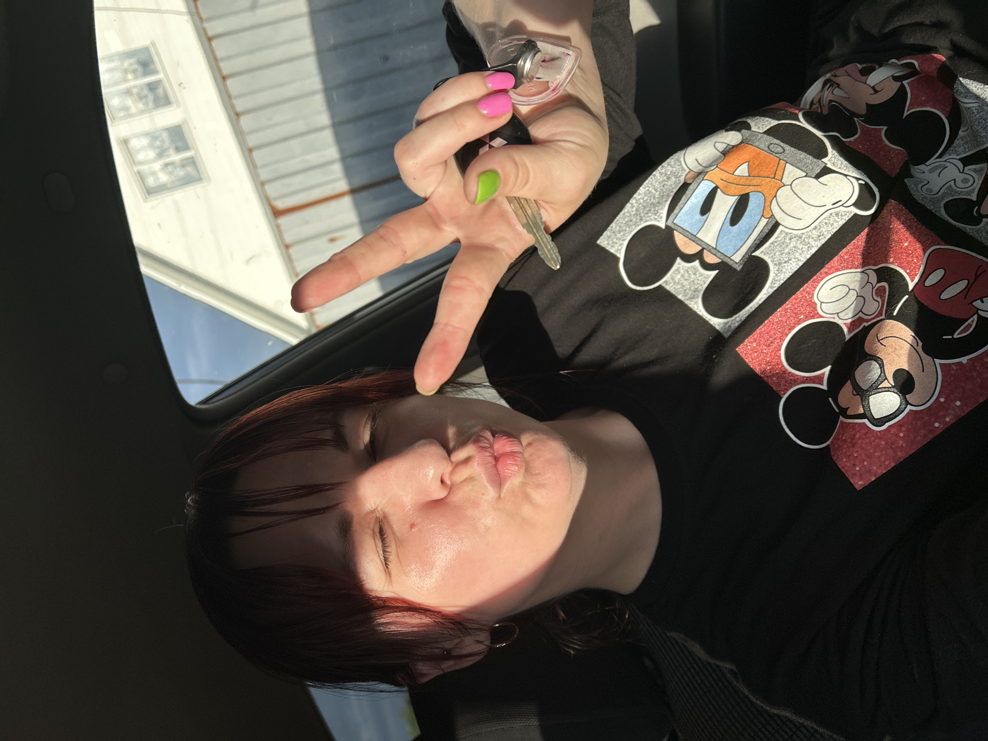 The image depicts a young woman with reddish-brown hair and bangs, sitting in the driver's seat of a vehicle. She is leaning forward, puckering her lips in a kissing gesture, and has her eyes closed. She is wearing a black t-shirt with a graphic print of Mickey Mouse and Donald Duck. Her right hand is raised, with her index and middle fingers extended in a peace sign. She is holding car keys with a pink heart-shaped keychain in the same hand. Her fingernails are painted in bright colors: pink on her ring finger and thumb, and lime green on her index finger. The sunlight is casting strong shadows across her face, highlighting the right side of her face and neck. Through the car window, a white building with windows can be seen, along with a light blue sky. The context suggests this photo was taken outdoors in daylight, possibly in or near the city of Foky, Russia, given the mention of the city.
