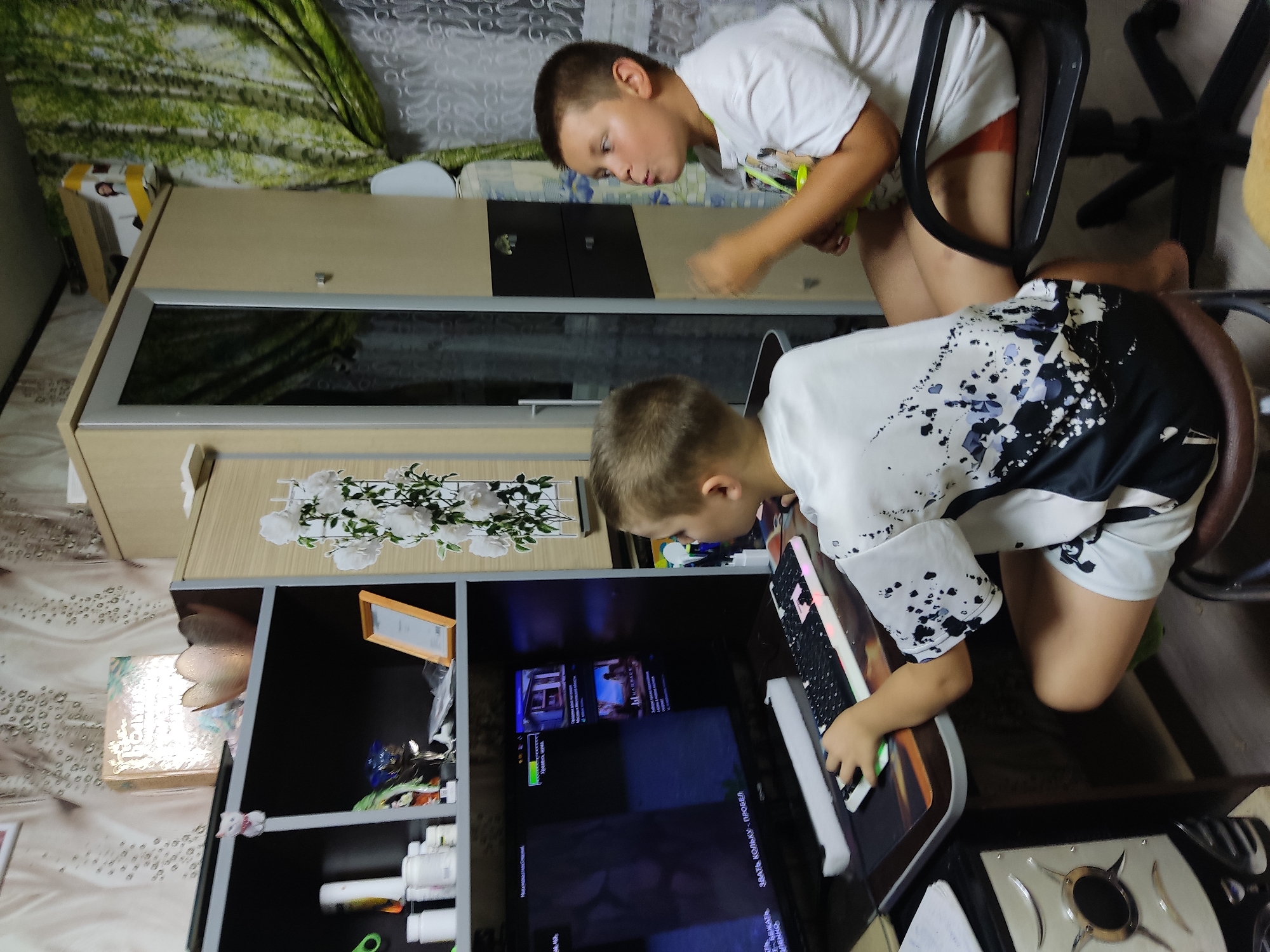 This image depicts an indoor scene, likely a residential setting, in Noginsk, Russia. Two young boys are engaged in an activity in front of a computer. The boy on the left, facing the computer screen, is wearing a white t-shirt with black and blue splatter-like graphics and white shorts. He is actively using a keyboard and mouse, suggesting he is playing a game or interacting with the computer. The computer screen displays a game interface with various menus and images. To the right, another boy, wearing a white t-shirt and white shorts with an orange band, is seated on a black office chair, looking towards the left. He appears to be holding a yellow object, possibly a snack or toy, and is engaged with the scene unfolding on the screen. The environment includes a desk with the computer setup, shelves with various items including decorative objects, books, and other miscellaneous items. Behind the desk is a tall cabinet with mirrored doors, and to the right, a window covered by curtains featuring a nature-themed print of trees. The overall impression is of children spending leisure time at home.