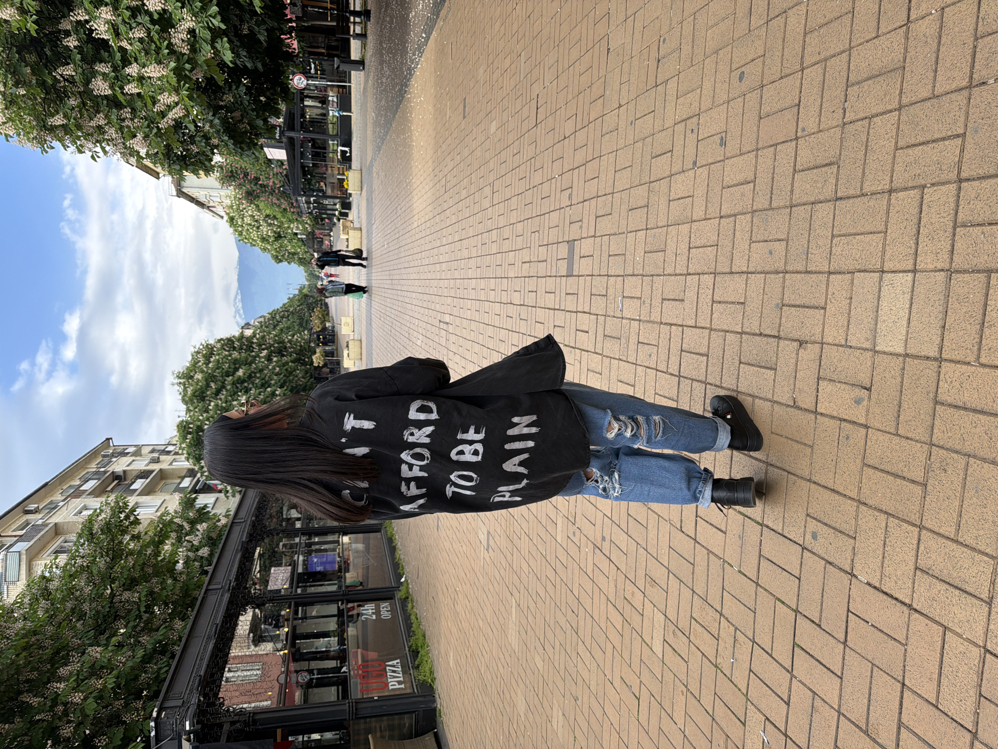 The image shows a person standing on a brick-paved street, wearing ripped blue jeans, black boots, and a black jacket with the words "I can't afford to be plain" written in white on the back. The person is facing away from the camera and has long, dark hair. The street is lined with trees and buildings. In the background, there is a view of mountains. A sign indicates that it is in Sofia, Bulgaria. A Pizza restaurant is visible to the left. There are other people walking in the distance. The sky is blue with some clouds.