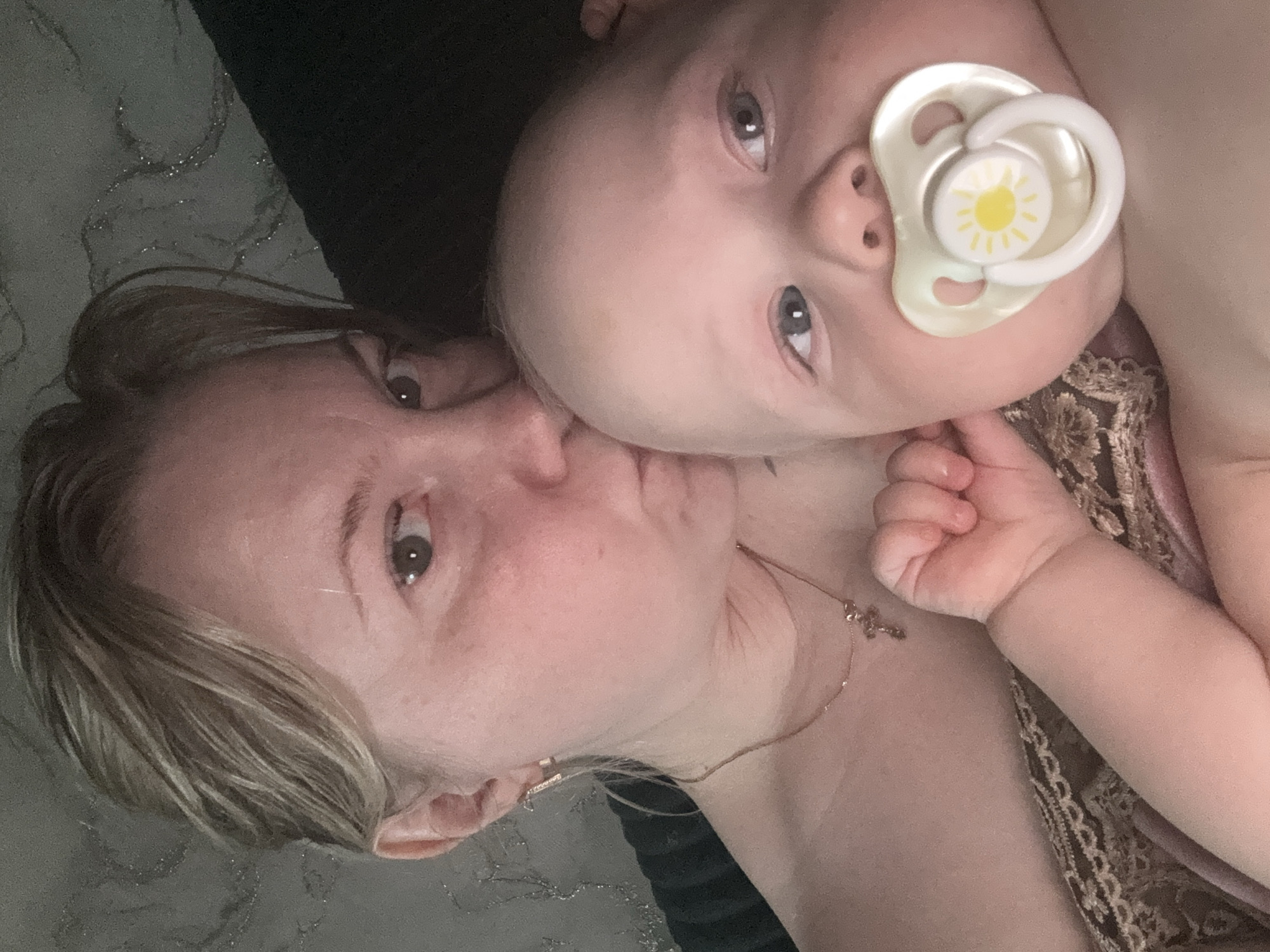 An intimate, close-up photograph features a woman and a baby lying together, possibly indoors in Chaikovsky, Russia. The image is taken from a slightly diagonal, overhead perspective.
The woman, with light skin and light-colored hair, is positioned on the left, looking towards the right side of the frame. Her eye, visible on the left, appears brown. Her lips are pursed, suggesting she is kissing the baby's cheek or temple, which is out of view but implied by her proximity. She wears a delicate necklace with a small cross pendant. A dark, possibly textured or patterned fabric, potentially a blanket or bedding, is visible behind her head and to her left.
The baby, also with light skin and light-colored hair, occupies the right side of the frame. Its face is turned towards the viewer, revealing bright blue or grey eyes. A white pacifier with a yellow sun design is in its mouth. A tiny hand is visible near the woman's neck/chest area, and the baby appears to be wearing light-colored clothing. Part of what appears to be lace or patterned fabric, likely part of the woman's attire, is visible beneath the baby's head and arm.
The background is dark and indistinct, suggesting a soft, possibly low-light environment. The overall mood conveyed is one of tenderness and affection between the two individuals. No specific location cues beyond the subjects are visible to suggest Chaikovsky, Russia. There is no visible text in the image.