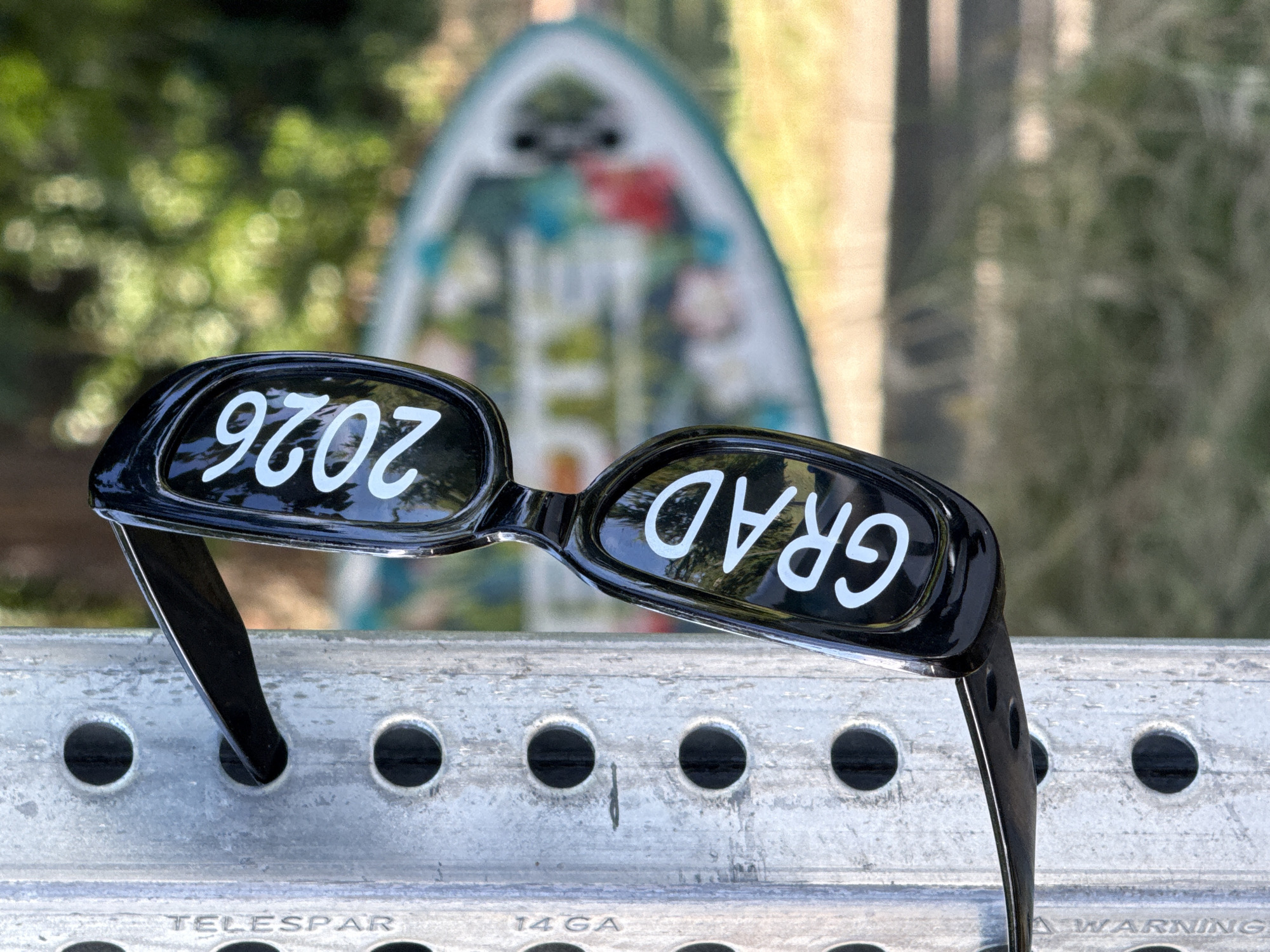 A close-up, eye-level, outdoor medium shot shows a pair of black sunglasses with "GRAD 2026" written in white text on the lenses. The sunglasses are hanging from a silver metal pole that has multiple circular holes. The pole has text on it, including "TELESAR" and "14 GA". In the background, a blurry green and yellow environment suggests trees and grass. There is also a blurred, colorful object, possibly a surfboard or a kayak, in the mid-background. The overall lighting suggests a sunny day.