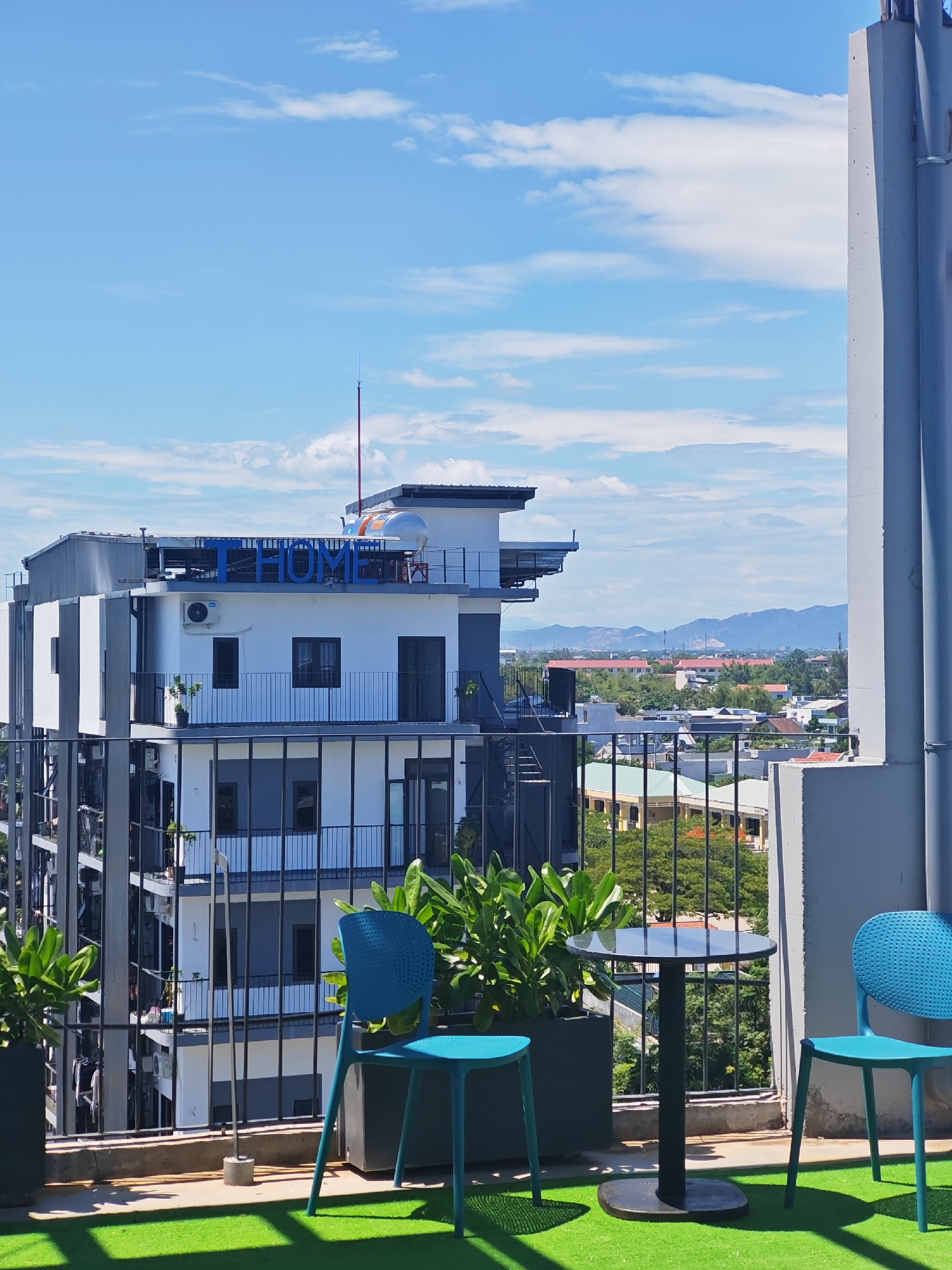 An outdoor seating area with artificial grass and a black circular table is on a balcony overlooking a city. Two blue chairs are positioned at the table. In the background, a modern white building with blue "T HOME" signage stands against a blue sky with scattered clouds. The building has multiple balconies and air conditioning units visible. Beyond the building, a sprawling urban landscape with various structures and lush green trees stretches towards a range of distant mountains. The scene appears to be a rooftop or elevated balcony in Đà Nẵng, Vietnam, offering a view of the city and its surrounding natural environment.