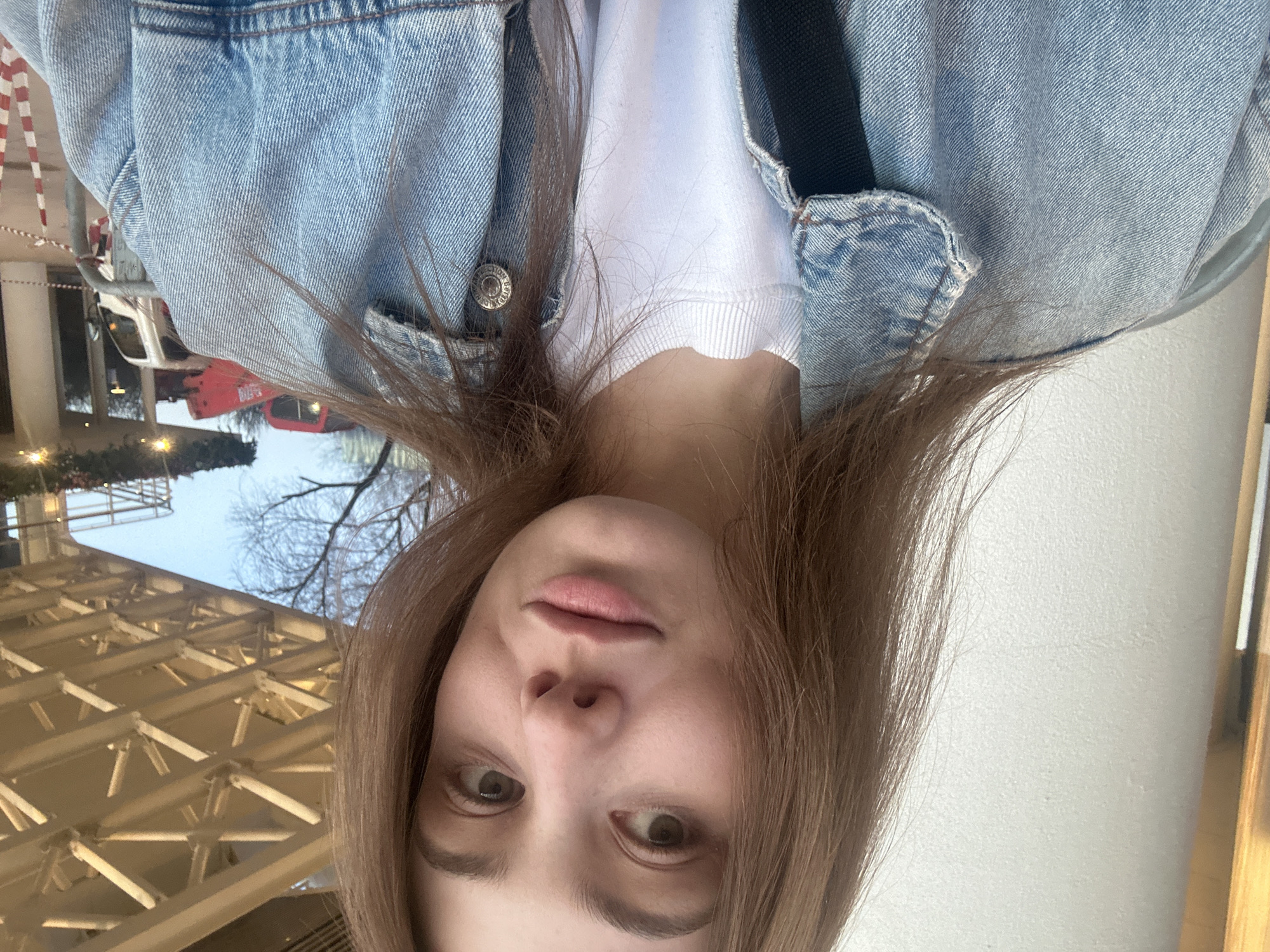 A selfie captures a young woman with brown hair, light skin, and neutral expression. She is wearing a white t-shirt and a light blue denim jacket with a button visible. A black strap is draped over her shoulder.
The background features an outdoor scene. To the left, there's a structural element with a grid-like pattern of light-colored beams. Beyond this structure, a street scene is visible, including a red vehicle and other elements suggesting an urban environment.
The location appears to be Moscow, Russia, based on architectural and environmental cues consistent with the city's design and atmosphere.