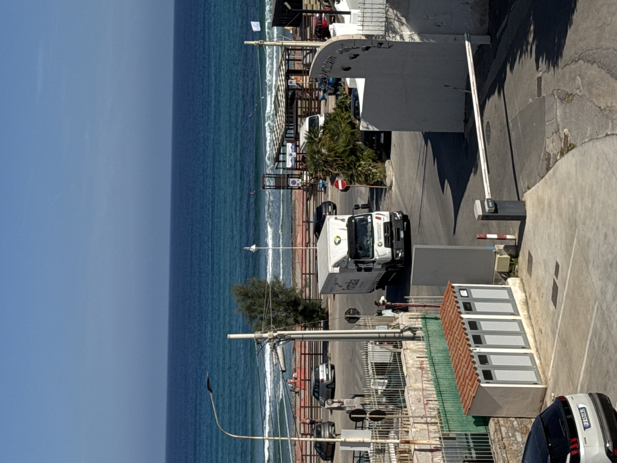 The image depicts a vibrant coastal scene under bright, clear skies. Although the image is oriented vertically with the sky on the left, the scene, if viewed upright, shows the sea stretching across the mid-ground, meeting a paved road and buildings on the right.
The sky, a brilliant blue, occupies the upper left portion of the image. It meets the turquoise sea, which is characterized by gentle white waves breaking along the shore. Several individuals can be seen scattered along the beach or shoreline, enjoying the sunny weather.
A paved road or parking area runs parallel to the coast. A white box truck, prominently featured, appears to have "Formaica" printed on its side and a "BOSCH" logo on its cab. Another white car, with an Italian license plate "EF 100TJ", is partially visible in the bottom right corner. Other vehicles are also parked or present along the road.
The land area is populated with various structures, including concrete walls and buildings. A white barrier gate is visible, controlling access to a paved area that features a red and white "no entry" sign. A distinctive long, low building with a red tiled roof and multiple gray-doored compartments stands out. Palm trees and other green foliage are interspersed among the buildings, providing shade and natural elements. Several streetlights or utility poles are also visible.
Visible text includes "Formaica" and "BOSCH" on the truck, "EF 100TJ" on a car's license plate, and fragments like "MANDARIN S..." and "LIDO..." on signs near the beach, suggesting seaside establishments. The bright illumination indicates it is mid-day, with sunny weather prevailing. The scene captures a lively, urbanized coastal setting, likely in Capaci, Italy.
