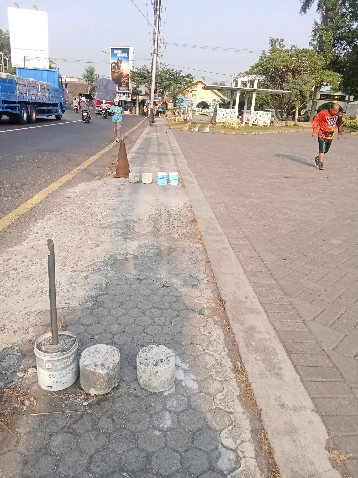 The image depicts a street scene in Kaliwungu, Indonesia. A person in an orange shirt and black shorts is jogging on a paved walkway. In the background, several people are visible near buildings and a billboard advertising a motorcycle event. A blue truck and other vehicles are on the road. The foreground shows a concrete sidewalk with construction materials, including a metal pole in a paint bucket and two concrete cylinders. The overall environment appears to be a public area with a mix of residential and commercial elements.