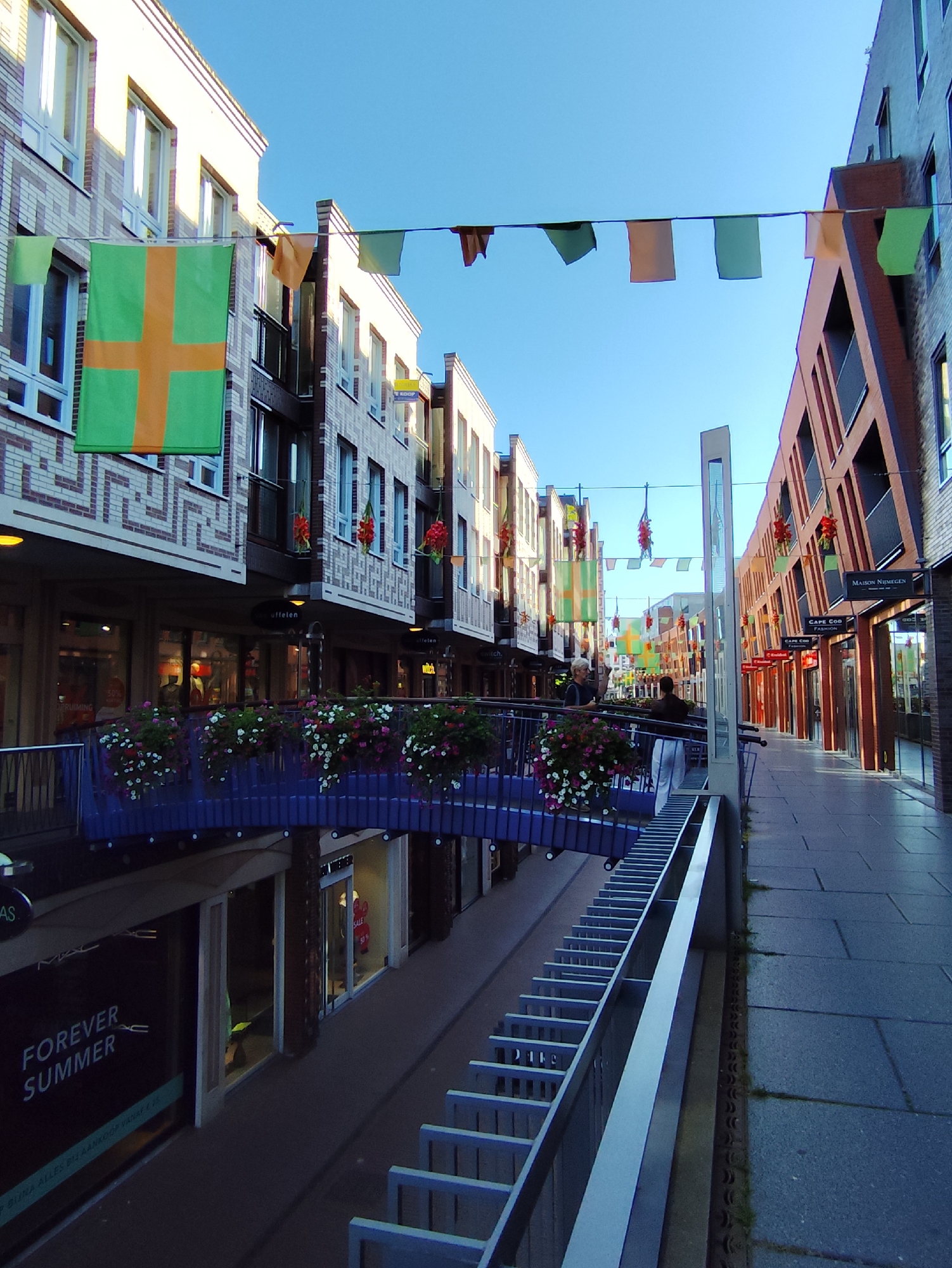 The image shows a street in Nijmegen, Netherlands, with buildings on both sides. The street is decorated with green and orange flags, and some red flower arrangements hang from the buildings. There are people walking on the street and on a bridge that crosses over the street. The buildings are modern with brick and glass facades. The street is paved with stone. There is a store with a sign that says "FOREVER SUMMER."