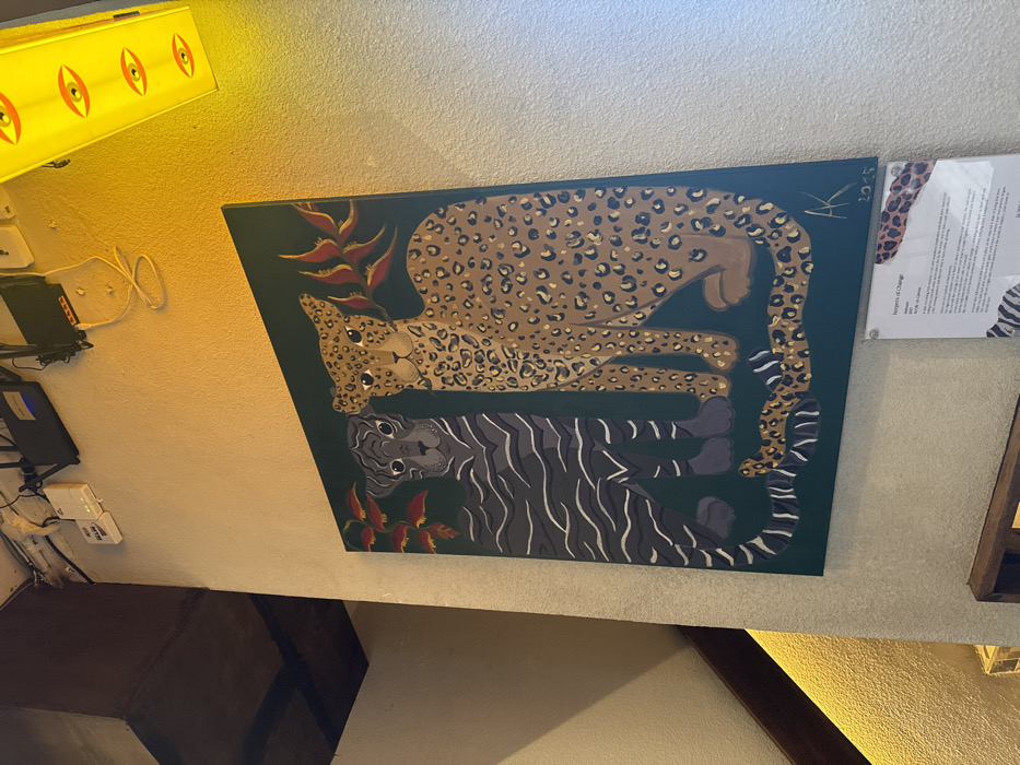 The image captures an interior scene featuring a prominent painting on a light-colored, textured wall. The painting is rectangular, oriented horizontally, and depicts two large feline creatures against a dark green background.
The upper animal is a leopard or jaguar, painted in shades of tan and brown with numerous black rosettes (spots). It is depicted lying down, facing left, with its head slightly raised and looking forward. Its eyes are dark, and its mouth is closed. Its tail curls towards the right side of the canvas.
Below the leopard, and mirroring its pose, is a tiger. This animal is rendered in dark grey with distinct black stripes. It is also lying down, but faces right, with its head raised and looking forward. Its eyes are dark, and its tail curls towards the left side of the canvas.
Stylized plant elements, possibly tropical flowers or leaves in shades of reddish-brown, are interspersed in the dark green background. One cluster appears near the leopard's head on the upper left, and another near the tiger's tail on the lower right. In the top right corner of the painting, a signature "AK" and the date "2023" are subtly visible.
The wall itself is a light, creamy off-white or beige color with a textured finish.
Above and to the left of the painting, a long, rectangular overhead light fixture emits a warm yellow glow. This fixture has several circular or oval cutouts that resemble eyes.
Further to the left, below the light fixture, several electronic devices, including black boxes and coiled white cables, are mounted on the wall. A white power strip is also visible.
To the right of the painting, an information sign is displayed in a transparent acrylic frame. The paper within the frame features abstract animal print patterns (leopard spots and zebra stripes) along its edges. The title "Natures of Change" is clearly readable at the top of the sign. Below the title, a paragraph of text provides context for the artwork, explaining that the leopard and tiger represent "strength, adaptability, and the interconnectedness of all living things," with the leopard's spots symbolizing "individuality and unique experiences," and the tiger's stripes signifying "unity and collective action."
In the lower part of the image, a dark brown, possibly wooden, rectangular object (like a piece of furniture or a shelf) is visible, along with a darker brown wooden beam or trim running diagonally.
The overall lighting in the scene is warm and bright, originating predominantly from the yellow light fixture, creating a cozy atmosphere.