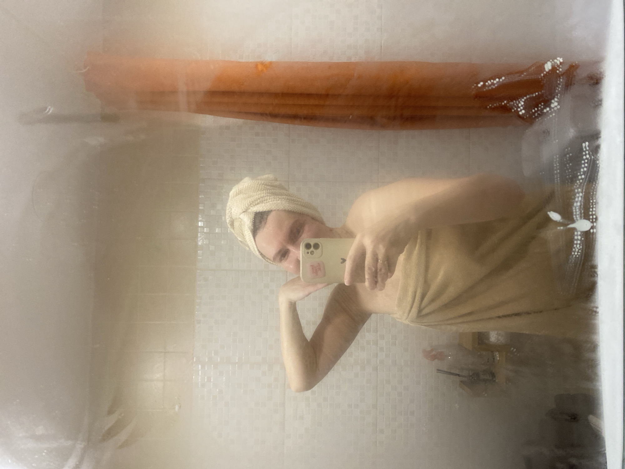 A person, wrapped in a beige towel and with a beige towel on their head, is taking a selfie in a steamy bathroom. The person is holding a phone with a light-colored case and a small sticker. Behind the person is a tile wall. A blurry shower curtain is visible, along with some bathroom accessories on a shelf. The location is likely Chaikovsky, Russia.