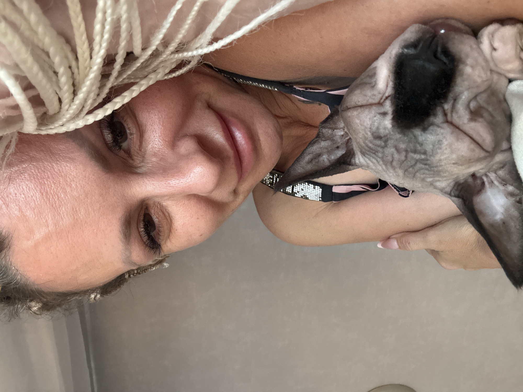 A close-up, eye-level shot shows a woman with braided blonde hair and a sphinx cat lying down. The woman is wearing a black bikini top with bejeweled straps. Her left arm is bent, and her hand rests on her chest, while the cat is nestled against her. The cat is grey with darker markings around its eyes and nose. The background is a plain, light-colored wall.