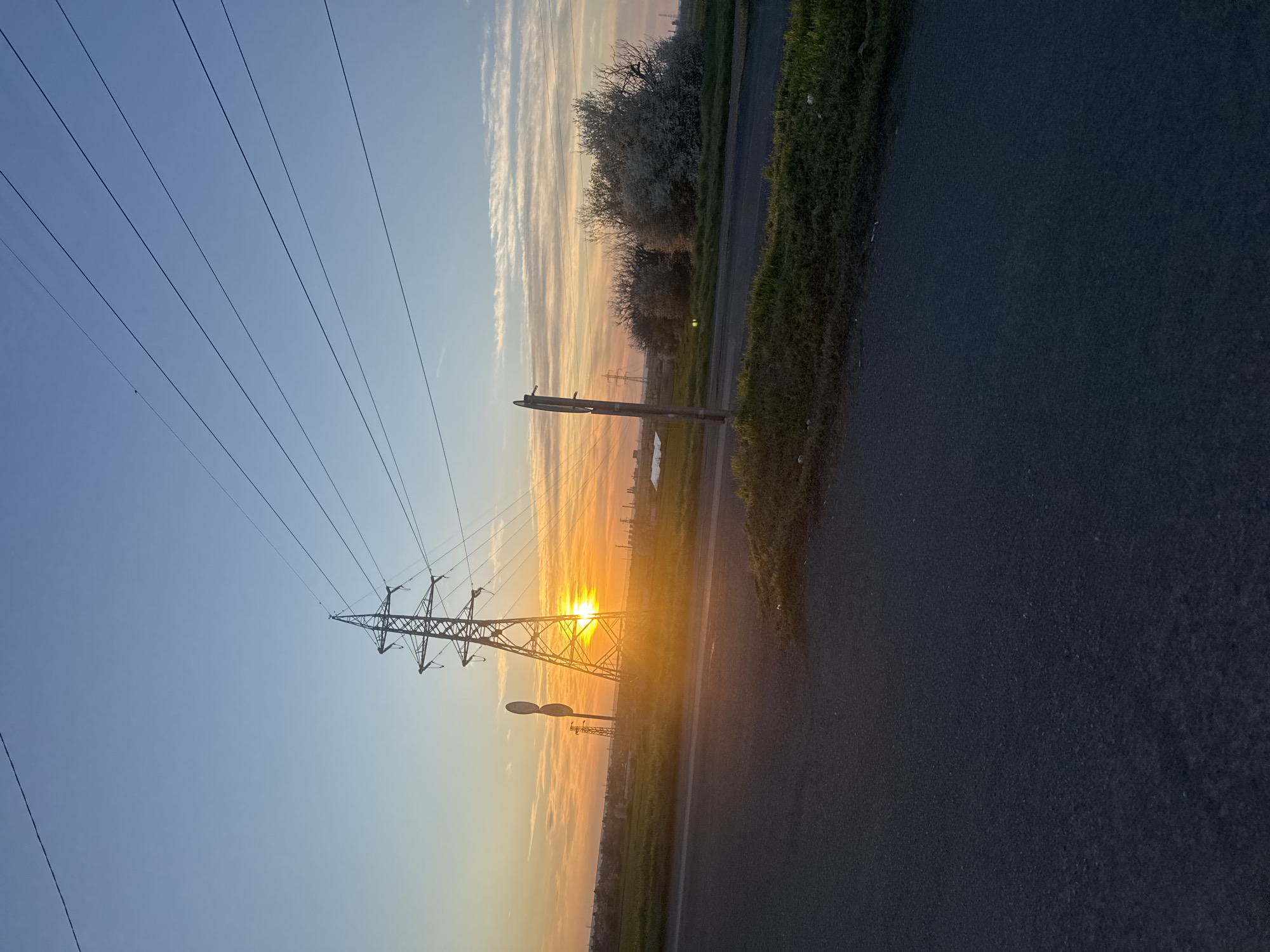 The image depicts a view at sunrise or sunset, showcasing a high-voltage transmission tower silhouetted against the bright light. Power lines stretch across the upper left of the frame. To the right of the tower, a road is visible, with vegetation lining its edge. In the background, trees and other structures are faintly discernible. There are no people visible in the image. There is no specific information to determine the precise location in Burgas, Bulgaria.