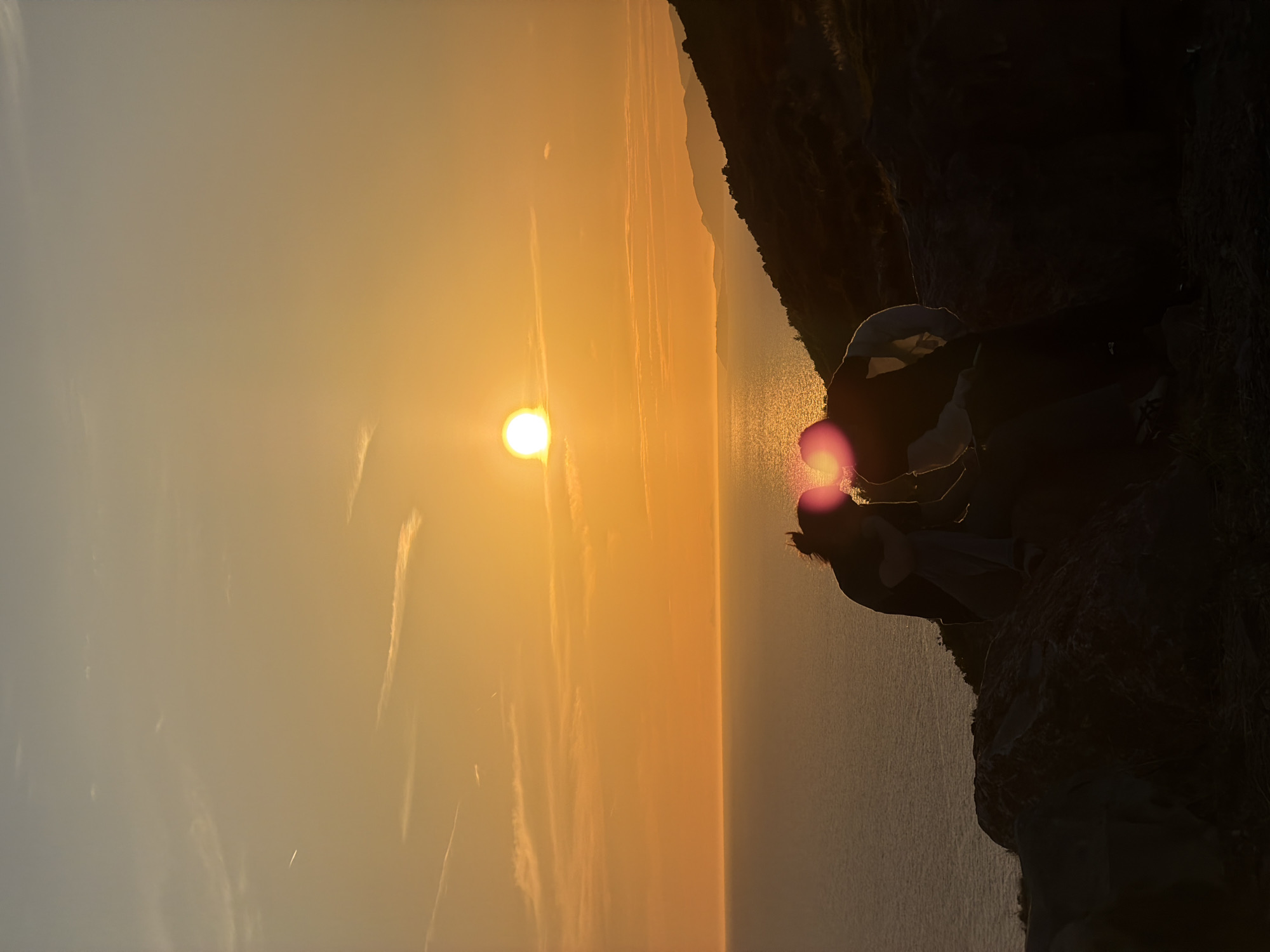 A silhouette of two individuals is captured in the foreground, sitting on a rocky outcrop overlooking the sea during sunset. The sun, a bright orb, is positioned in the center of the frame, casting a warm orange glow across the sky and water. The individuals are positioned close together, appearing to be sharing a moment. One individual, on the left, has their head bowed, with their hair tied up in a bun. The other individual, on the right, is leaning in towards the first. The sea stretches out to the horizon, reflecting the vibrant colors of the sunset. Hills with dark outlines are visible in the distance on the right side of the image. The overall scene depicts a tranquil and romantic natural setting.