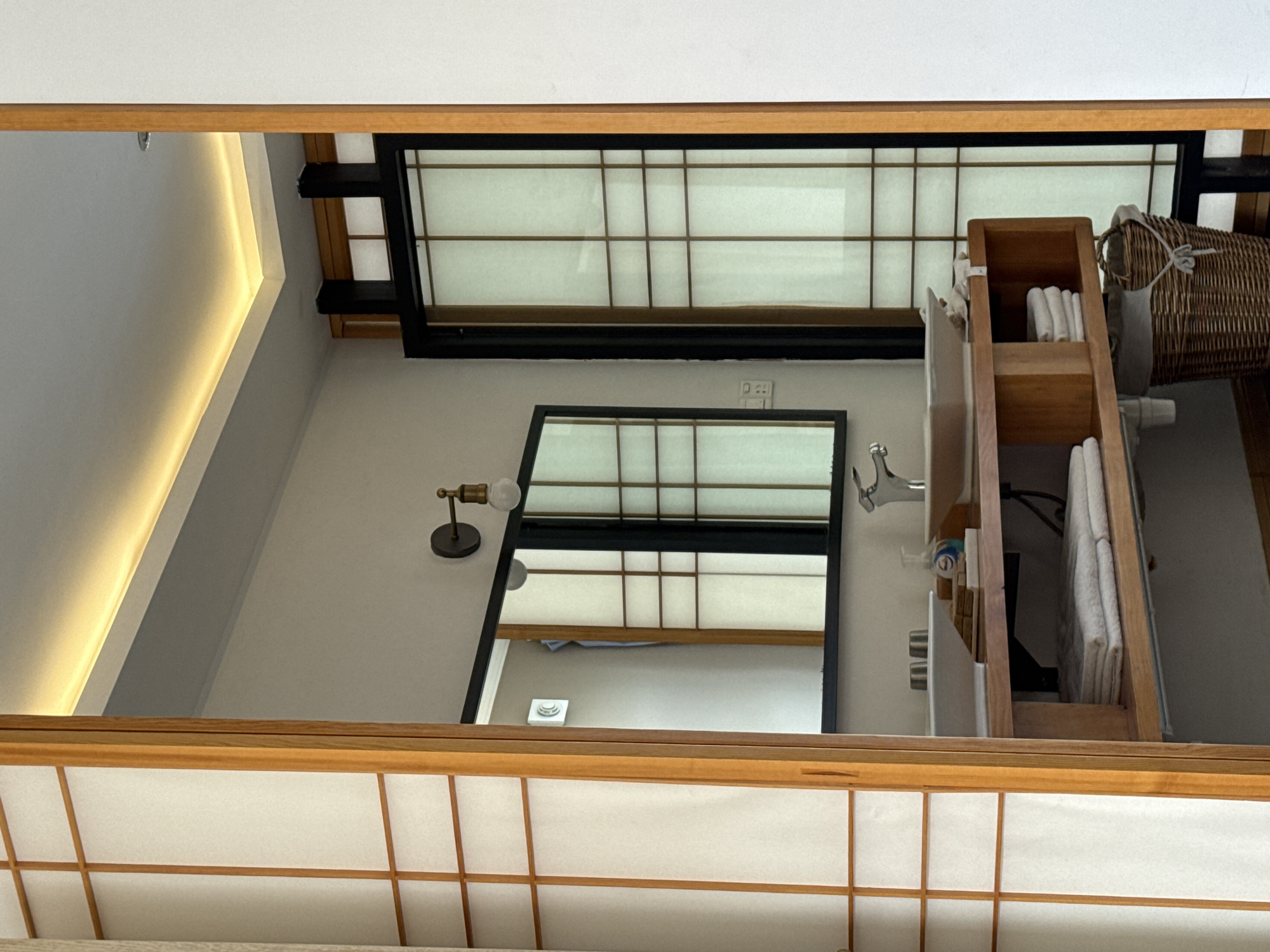 The image shows a modern bathroom with elements of traditional Japanese design, located in Dali City, China. The bathroom features a wooden vanity with a white sink, a chrome faucet, and several stacks of neatly folded white towels. To the right of the sink, a woven laundry basket is visible. The vanity is framed by wooden elements and a white wall. Above the vanity, a large mirror reflects a wall with a light fixture and a sliding door with a Shoji screen design, which is also visible to the right of the vanity. The ceiling has subtle, warm LED lighting. The overall impression is one of clean, minimalist design with natural materials. There are no people present in the image.