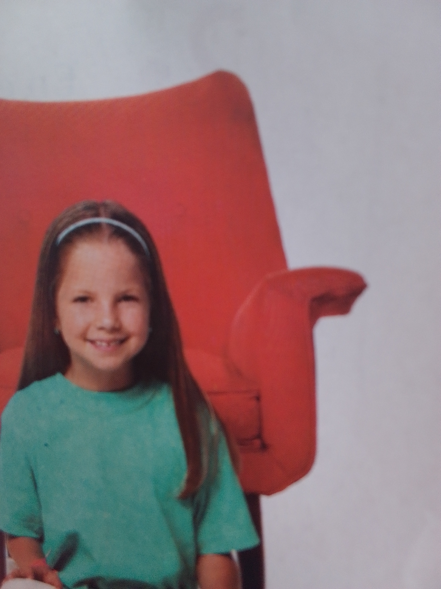 A young girl with long brown hair and a blue headband is sitting in a large red armchair. She is wearing a turquoise t-shirt. The background is plain white. This image appears to be a studio portrait, not depicting any specific location in La Tagua, Venezuela.
