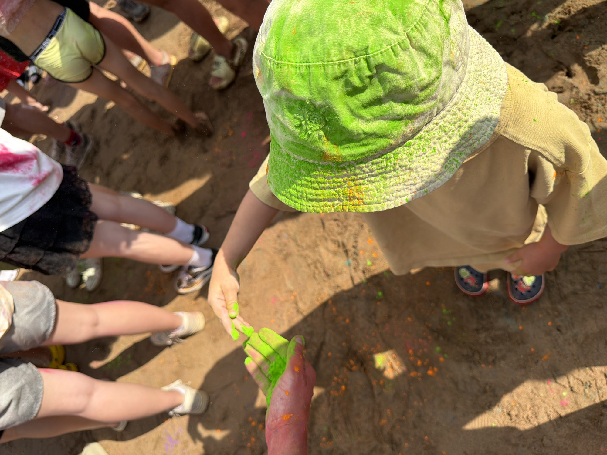 In Chaikovsky, Russia, a group of people, primarily young individuals, are participating in a color powder festival, likely at an outdoor sandy area, possibly a park or recreational ground. The ground is covered with scattered colorful powders, predominantly in shades of green, orange, and pink, indicating a vibrant and messy celebration.
In the foreground, two hands are prominently featured. One hand, appearing to belong to an adult or older youth, is holding a handful of bright green powder, which is being transferred to another hand. The recipient hand, belonging to a child, is open and cupped, ready to receive the powder. This child is wearing a light beige hooded sweatshirt and a green bucket hat, both liberally dusted with the colorful powders. Their feet are clad in dark sneakers with red accents.
Surrounding them, other participants are visible. Their lower legs and feet are shown, some wearing shorts, others wearing skirts. Their footwear includes sneakers and sandals. The legs of a person wearing yellow shorts are also visible. The overall atmosphere suggests a lively and energetic event, with participants actively engaging with the colored powders. The sunlight casts distinct shadows on the sandy ground, highlighting the texture of the sand and the scattered powder.