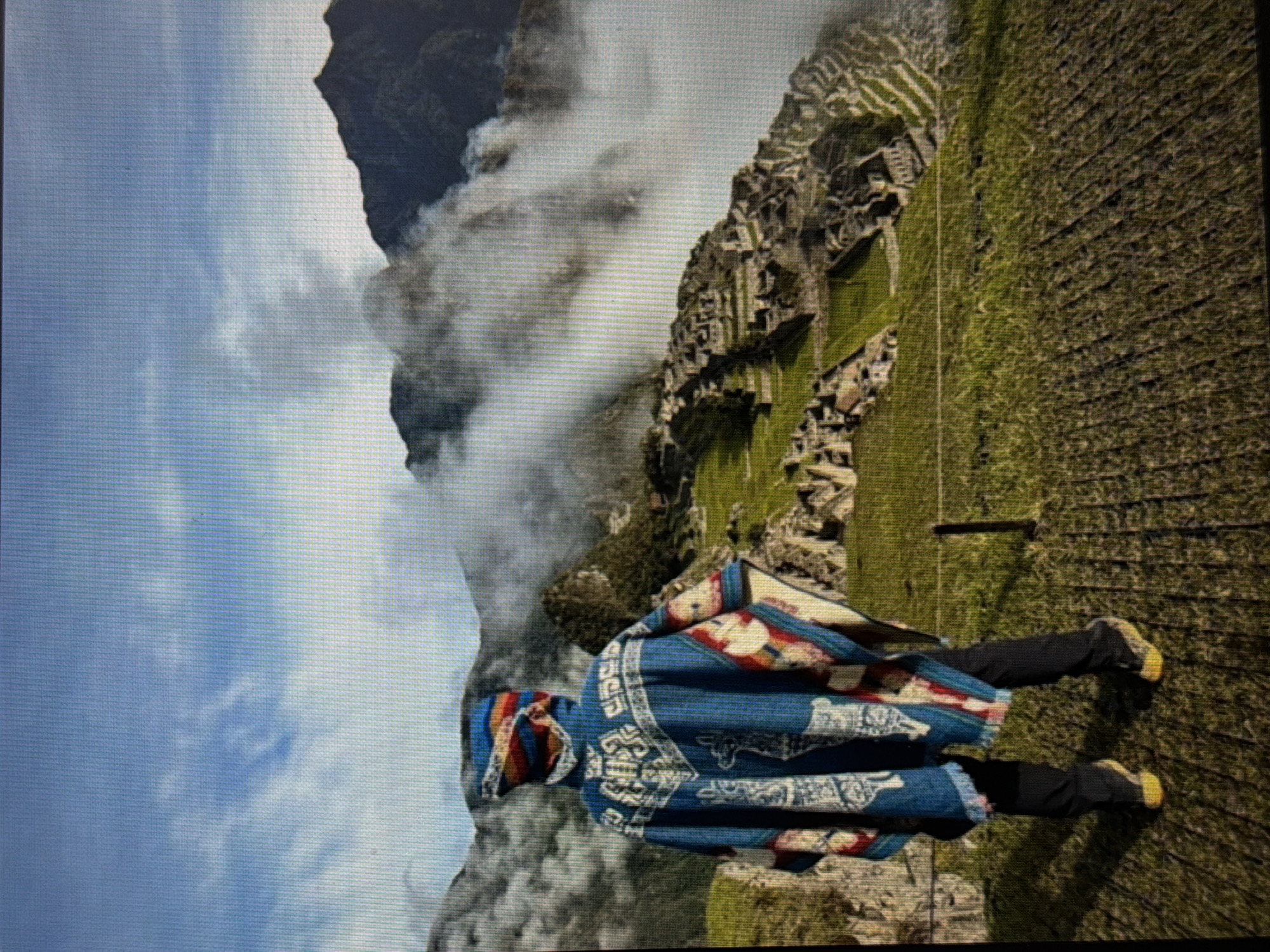 A person wearing a blue and red patterned poncho, black pants, and yellow sneakers stands in front of Machu Picchu in Peru. The sky is cloudy.