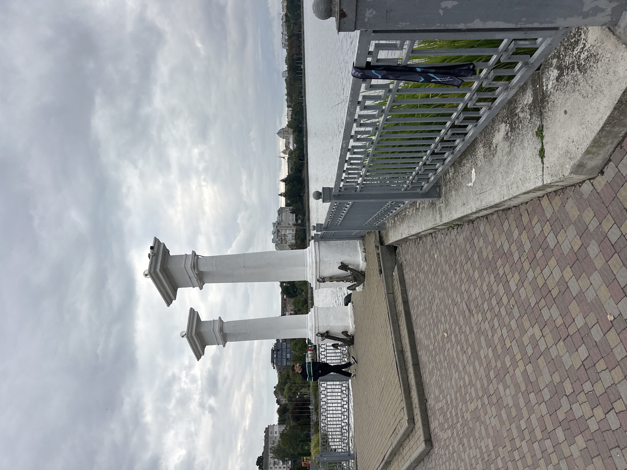 The image shows a man walking on a paved walkway next to a body of water in Ternopil, Ukraine. The walkway has two large white columns with anchors at their base. A grey metal fence runs along the edge of the water, with a dark item, possibly a jacket or bag, hanging from it. In the background, across the water, are buildings and trees. The sky is overcast. The location is likely the Ternopil Pond embankment.