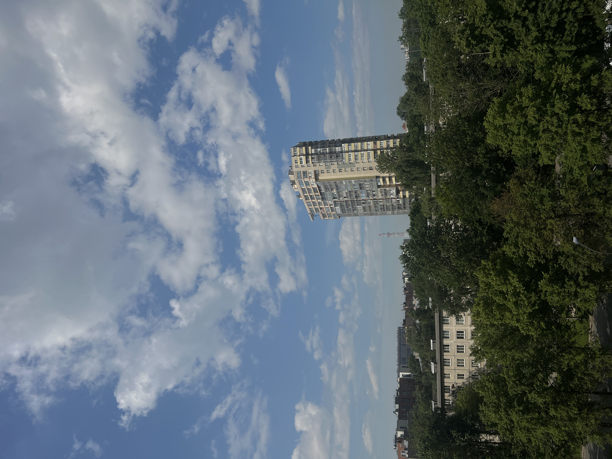 This is a daytime, exterior, high-angle shot capturing a cityscape in Petro-Slavyanka, Russia. The sky is predominantly blue with scattered, fluffy white clouds. In the background, a tall, modern apartment building with a cream-colored facade and numerous windows stands prominently. In the midground, a dense canopy of lush green trees covers a suburban area with several low-rise buildings peeking through the foliage. The overall impression is one of a quiet, residential urban environment bathed in sunlight.