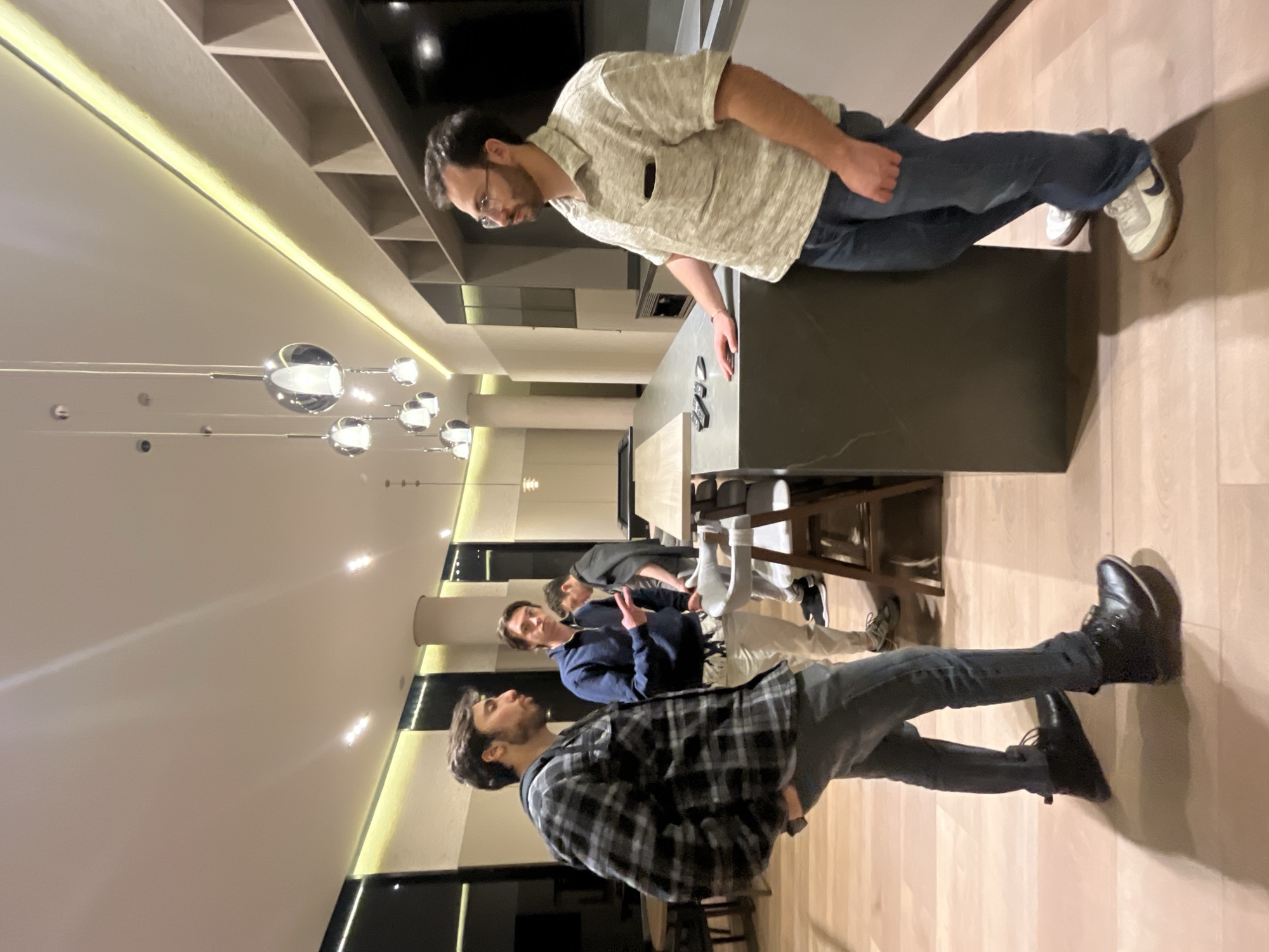 The scene depicts an interior view of a modern lounge or common area in Mexico City, Mexico, populated by four adult males. The flooring is light wood, complemented by a white ceiling featuring recessed lighting and multiple chrome spherical pendant lights. Linear LED strip lighting illuminates the upper sections of the walls, which integrate dark panels and light-colored shelving units.
In the foreground left, a man with dark hair and a beard, dressed in a dark grey plaid flannel shirt, dark jeans, and black boots, stands facing right with his right hand in a pocket. To his right, a second man in a navy blue half-zip sweatshirt and light-colored patterned pants stands facing the first man, gesturing with his right hand.
To the far right, a third man, wearing glasses, a light-textured short-sleeved polo shirt, and blue jeans with light-colored sneakers, leans against a dark grey stone-like counter. His left arm rests on the counter, adjacent to three dark-colored remote controls. A light wooden counter extension is visible behind this man, accompanied by bar stools.
In the background right, a fourth man is partially visible near a pool table, dressed in dark attire. Architectural features include a large circular pillar and a dark flat-screen monitor mounted above the main counter. A round table with chairs is located in the far-left background.