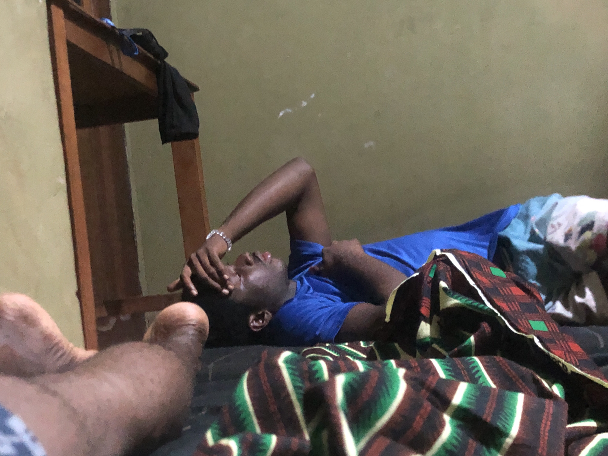 In Nwofia, Nigeria, a man is lying on a bed with a patterned blanket partially covering him. He is wearing a blue t-shirt and has his hand on his forehead. A wooden shelf stands beside the bed. Another person's feet are visible in the foreground. The background features a plain wall.