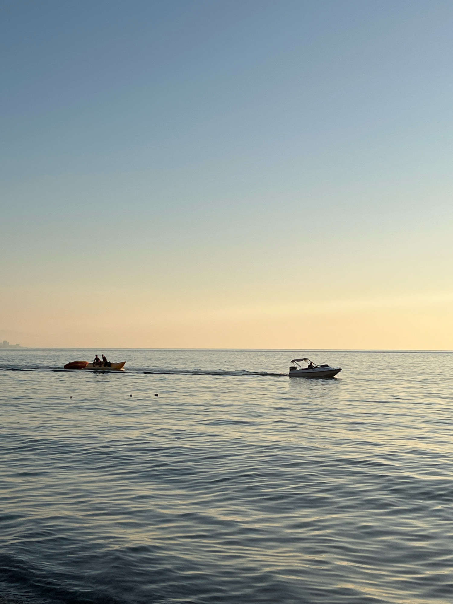 The image depicts a serene seascape at dusk, likely near Kobuleti, Georgia, given the distant city skyline visible on the left horizon. The sky transitions from a clear, light blue at the top to a soft, warm yellow gradient near the horizon, indicating the setting sun. The sea is calm, with gentle ripples reflecting the sky's colors.
In the foreground, two small boats are visible in the water. To the left, a yellow banana boat is being pulled by an unseen vessel, with two individuals seated on it. Further to the right, a white speedboat with a canopy is also moving through the water, with at least one person visible inside. A series of small, dark buoys or markers are spaced in a line across the water between the two visible boats, suggesting a designated area or a course. The overall scene conveys a sense of tranquility and leisure.
