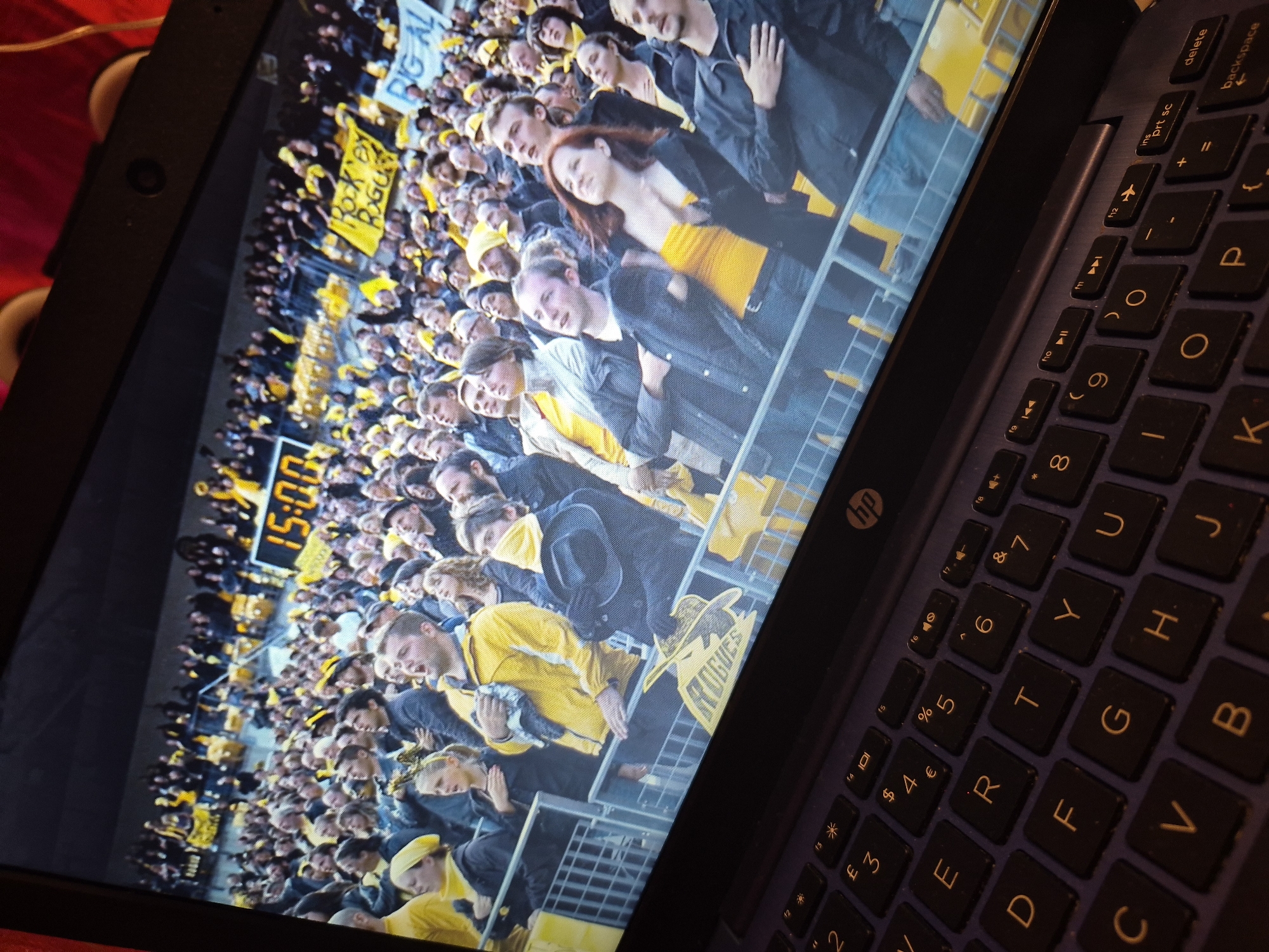 The image shows a laptop screen displaying a video of a large crowd of people at an outdoor event, possibly a sporting match or concert. The crowd is densely packed, and many individuals are wearing yellow clothing and hats. In the background, a large digital clock displays "15:00." There are also banners visible, one of which appears to read "ROCK BY ROGUE." The laptop itself is on a dark surface, with a keyboard visible in the foreground. The overall lighting suggests an indoor setting, likely a dimly lit room, with the laptop screen being the primary light source. The image does not provide any specific details that would allow for the identification of the city of Sandwell, United Kingdom.