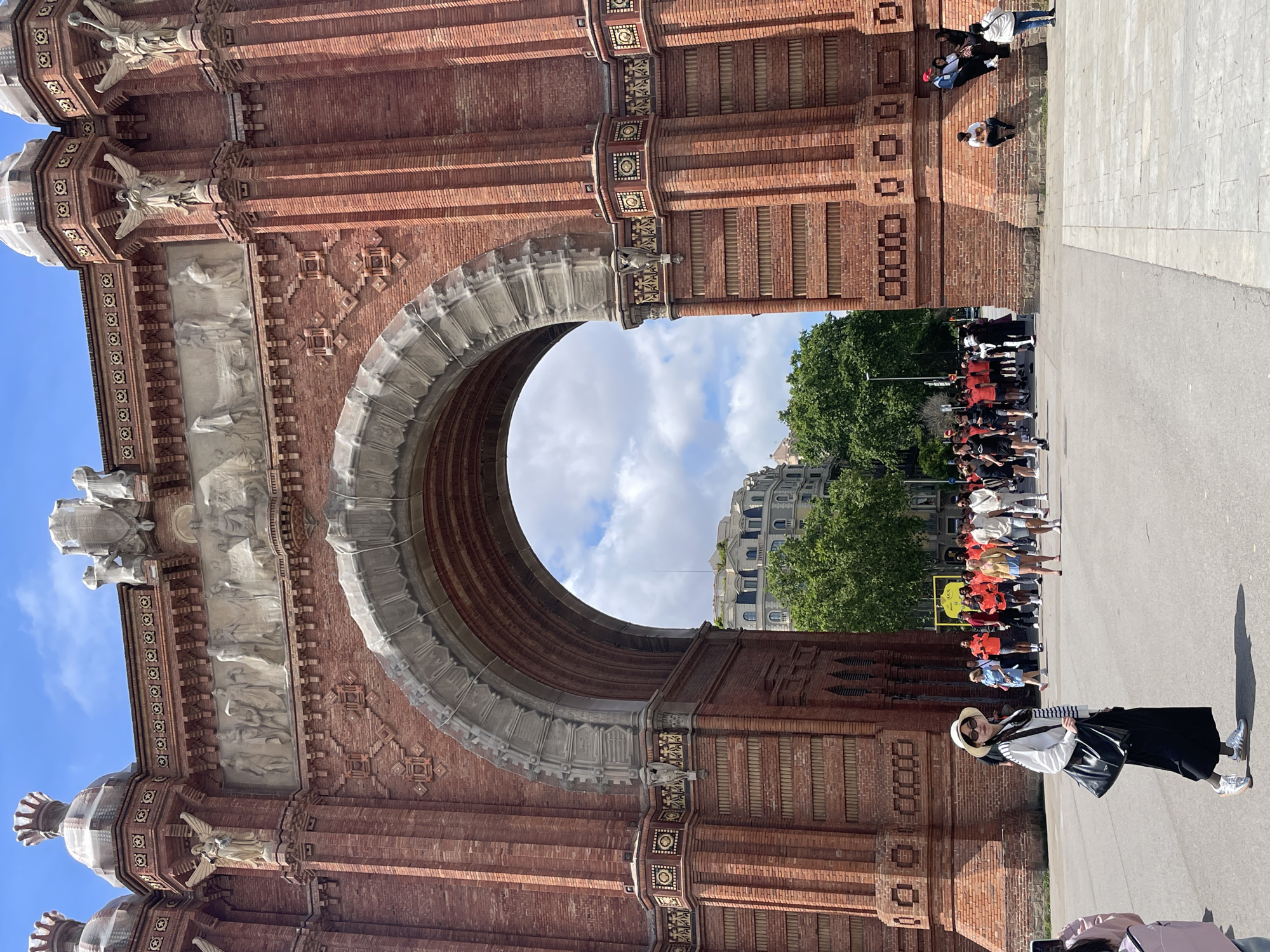 The image depicts the Arc de Triomf in Barcelona, Spain, from a ground-level perspective. The red brick arch, adorned with stone carvings and decorative elements, frames a view of buildings, trees, and a group of people. Some people are sitting on the brick wall of the arc. A person wearing a straw hat, striped shirt, and dark skirt is walking in front of the arch. The sky visible through the arch is partially cloudy. The photograph captures a sunny, outdoor scene with tourists and architectural features of Barcelona.