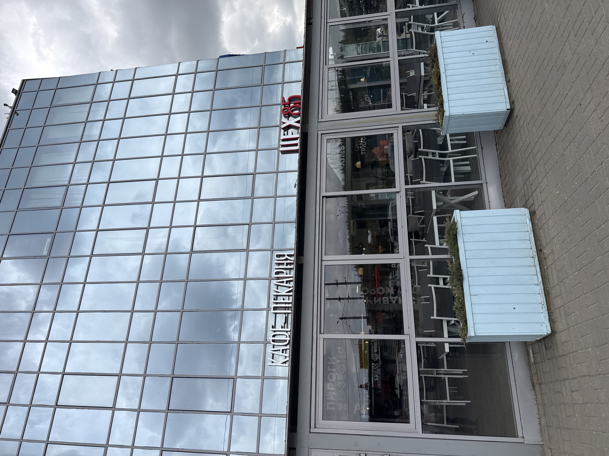 The image shows the exterior of a building located in Medovoye, Russia, which houses a cafe-bakery called "Shex85." The building has a modern architectural design with a glass facade that reflects the cloudy sky. The cafe-bakery's name is displayed on the building in both Russian and English. The cafe has a street-side seating area with chairs and tables visible through the large windows. Two light blue rectangular planters with greenery are located next to the seating area on the paved sidewalk. The overall impression is a modern and inviting cafe-bakery in an urban setting.